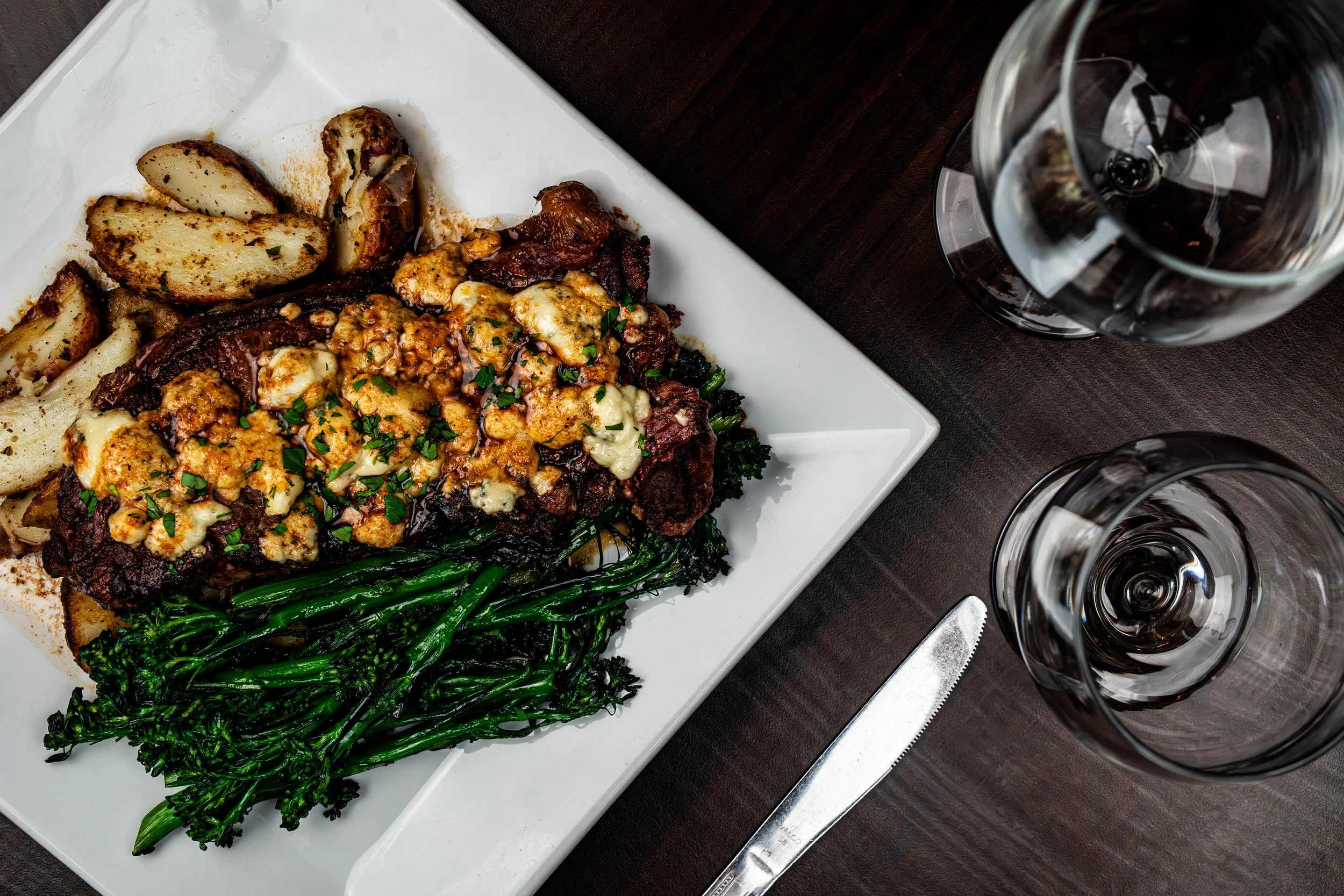A white square plate with a serving of roasted potatoes, cheesy beef casserole, and steamed broccolini on a dark wooden table, with two glasses of red wine and a knife nearby.
