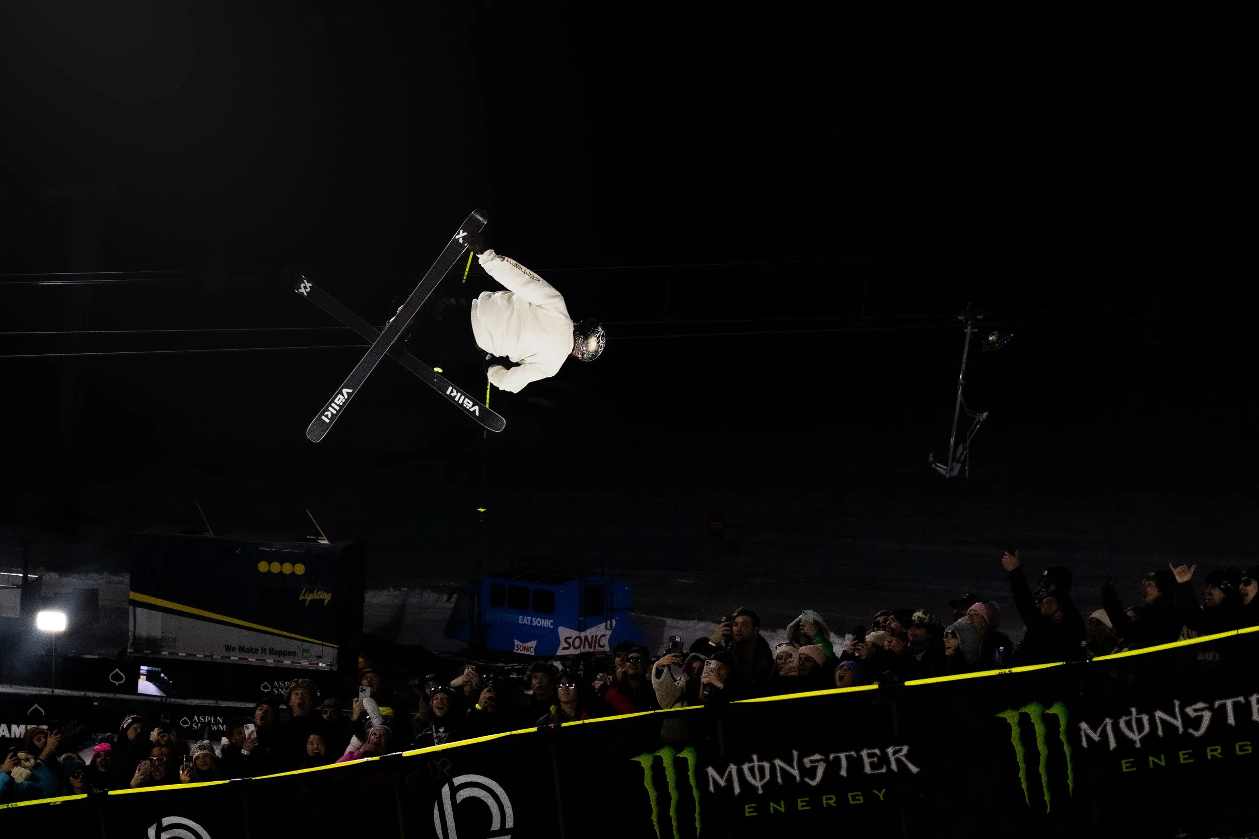 A skier in mid-air performing a jump during a night event with spectators watching and cheering in the foreground.