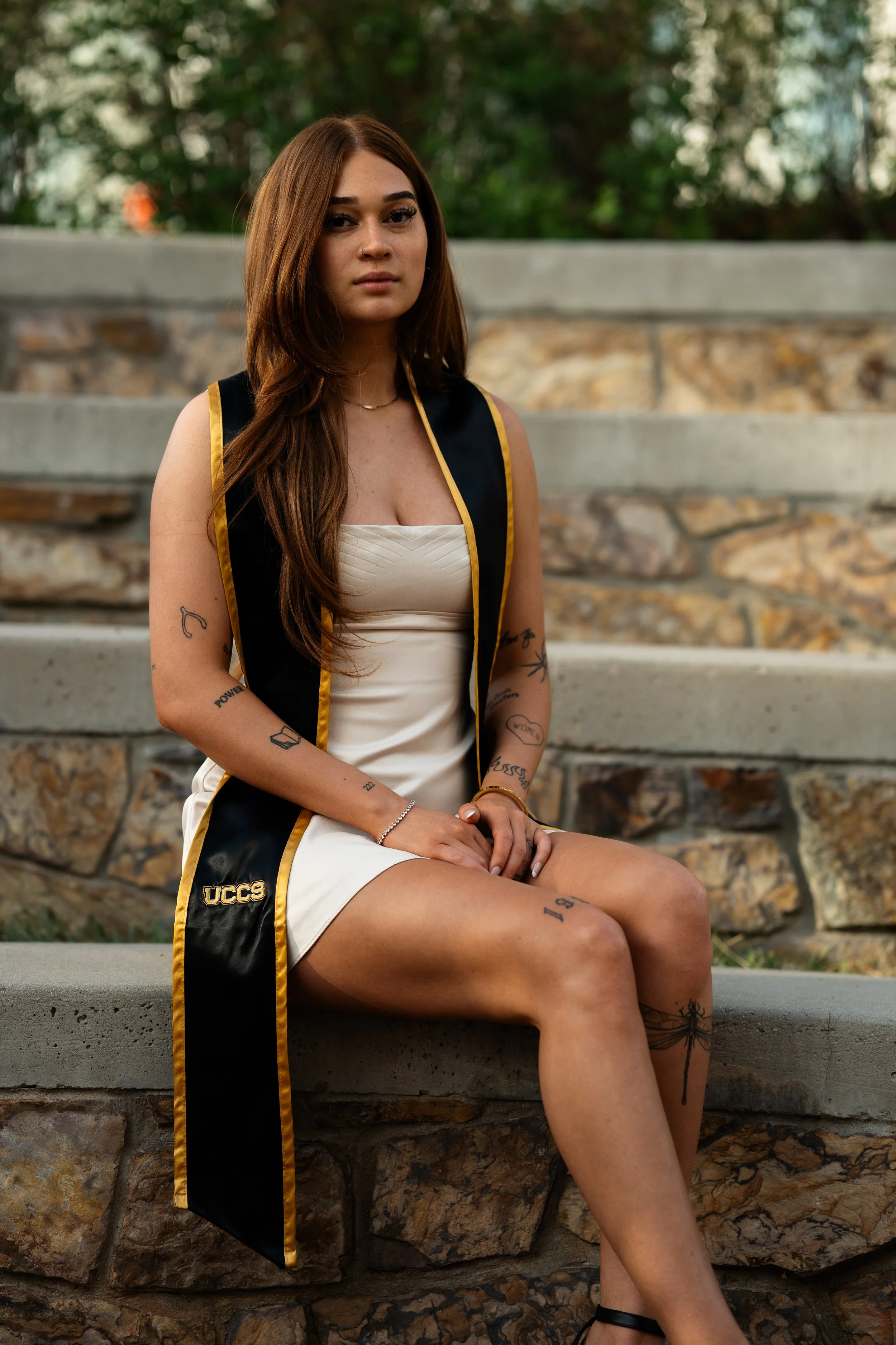 A young woman with long brown hair sitting on stone steps outdoors, wearing a cream dress and a black and gold graduation sash with "UCCS" on it, surrounded by greenery.