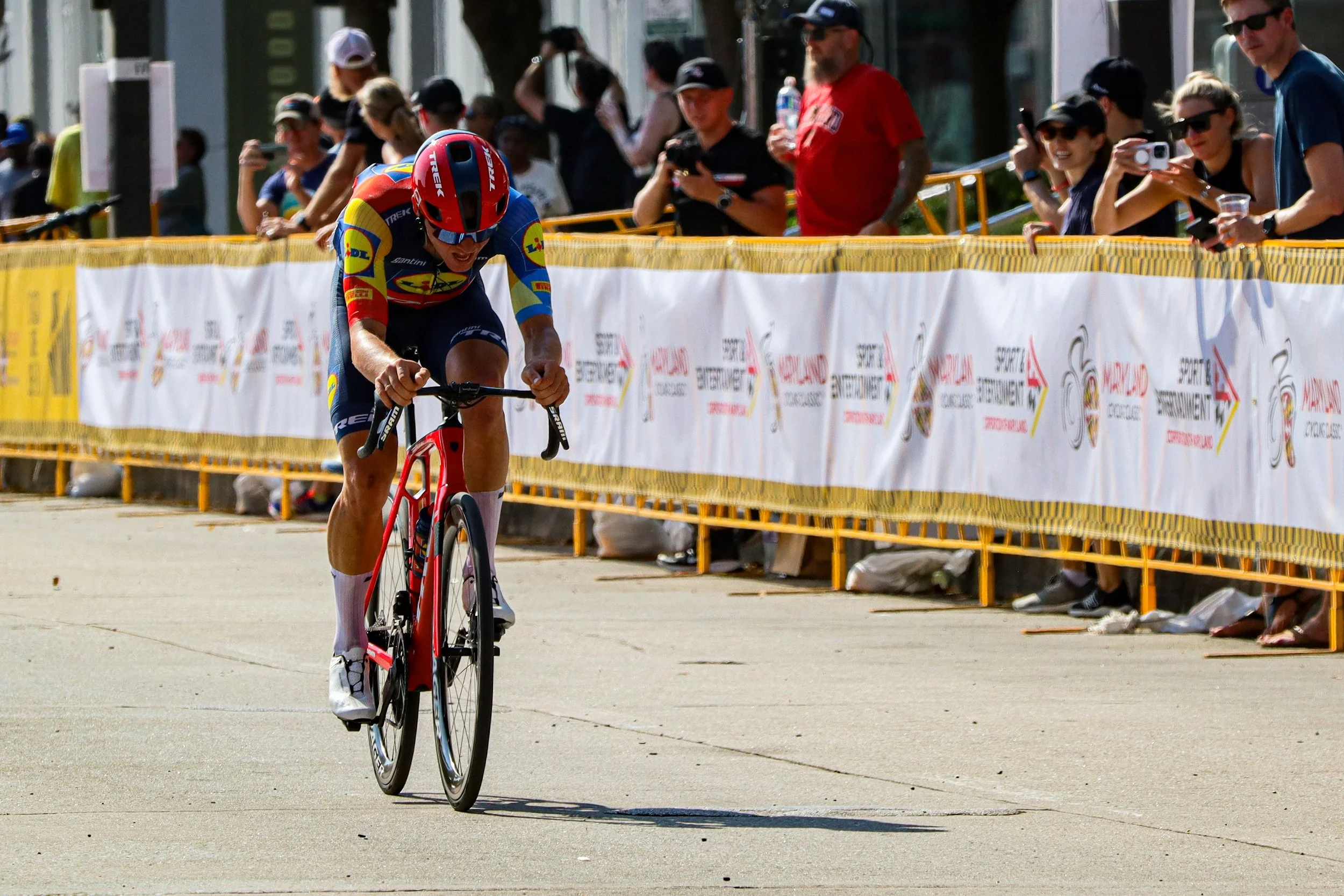 A male cyclist in a colorful cycling outfit and helmet competes in a race, riding a red bicycle on a paved street lined with yellow barriers, while spectators watch and photograph from behind the barriers.
