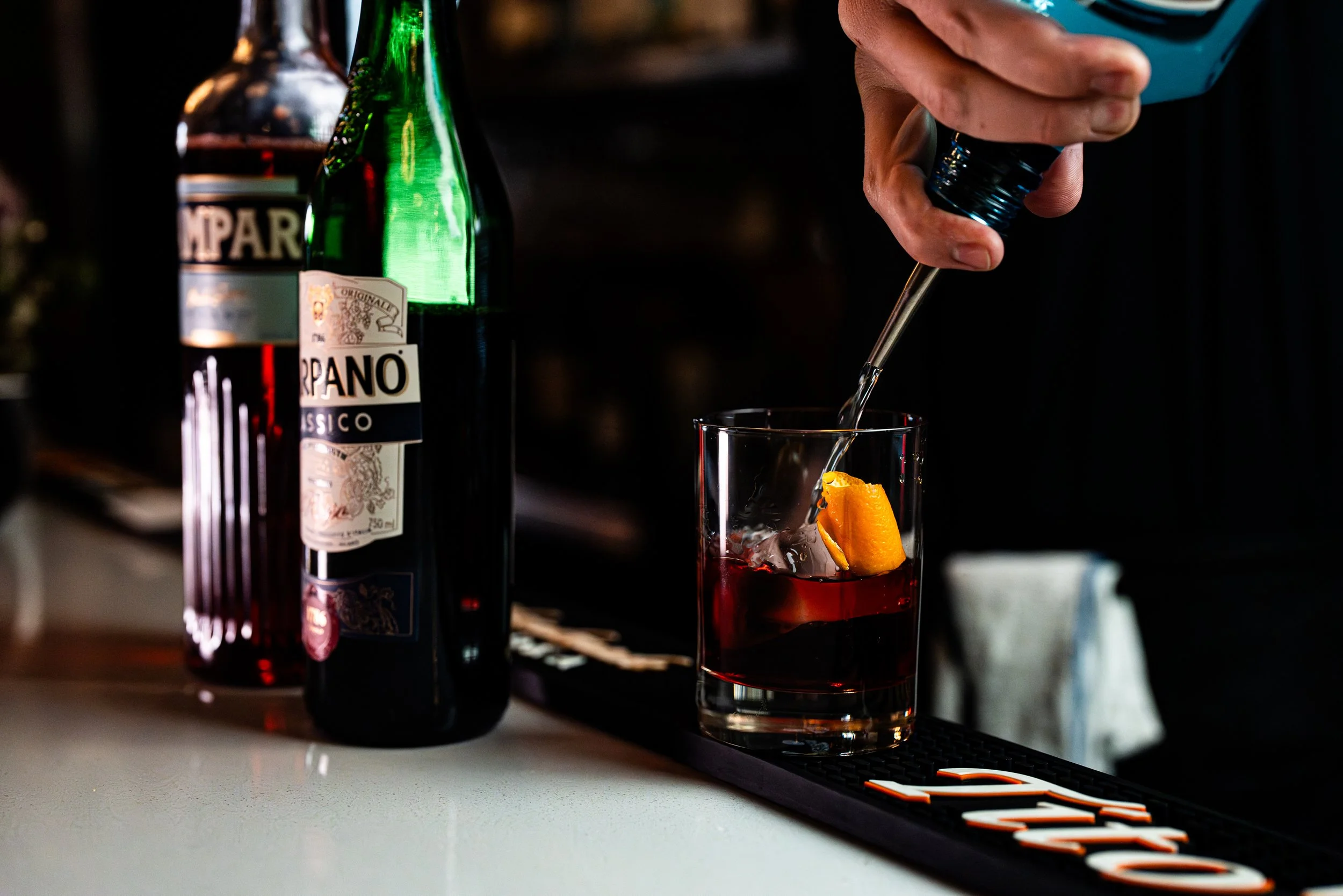 A bartender pours clear liquid into a glass with an orange slice, with bottles of alcohol and a bar mat visible on the counter.