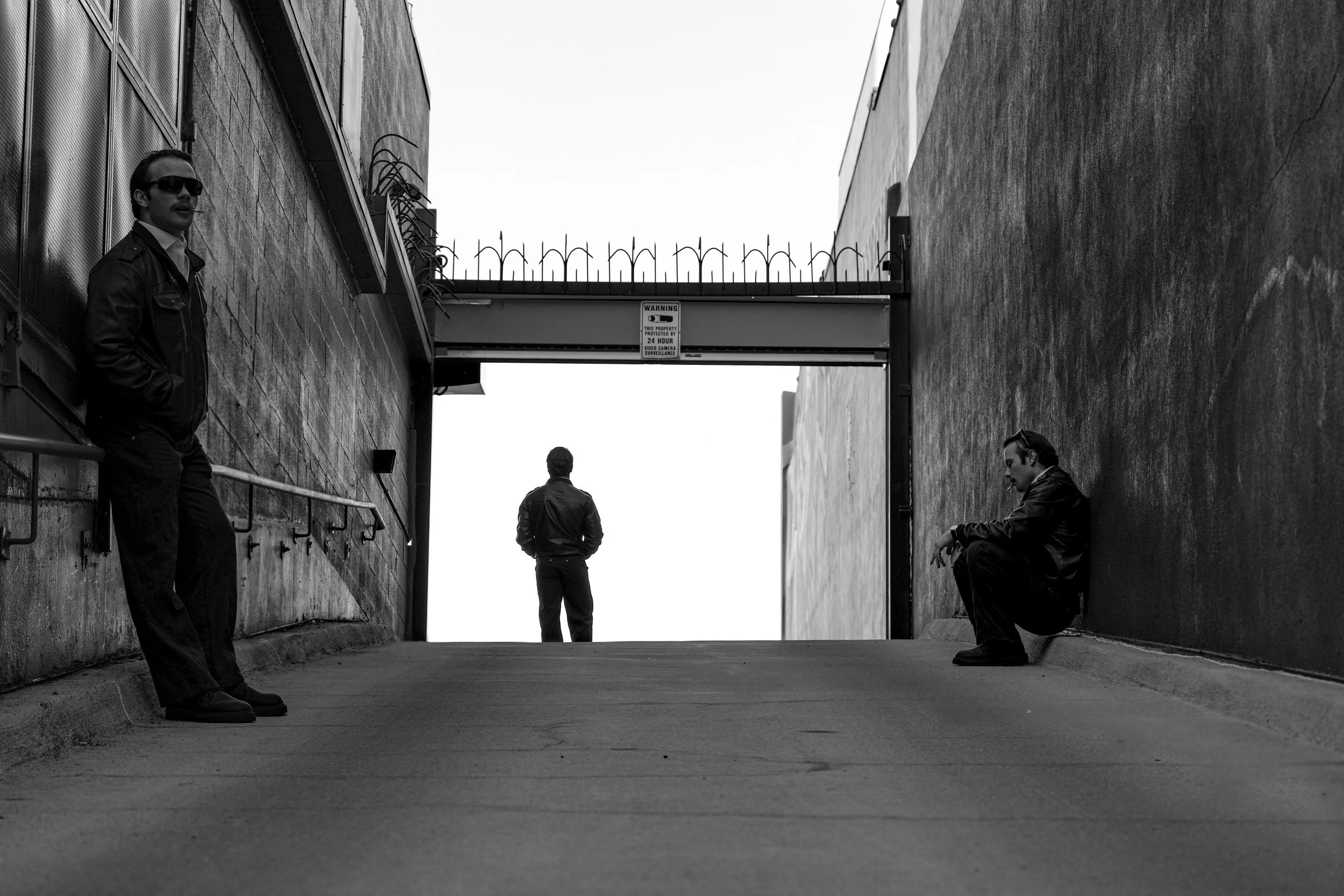 Three men in a dark alleyway, two sitting on the ground and one standing in the distance, with a bridge overhead, in black and white.