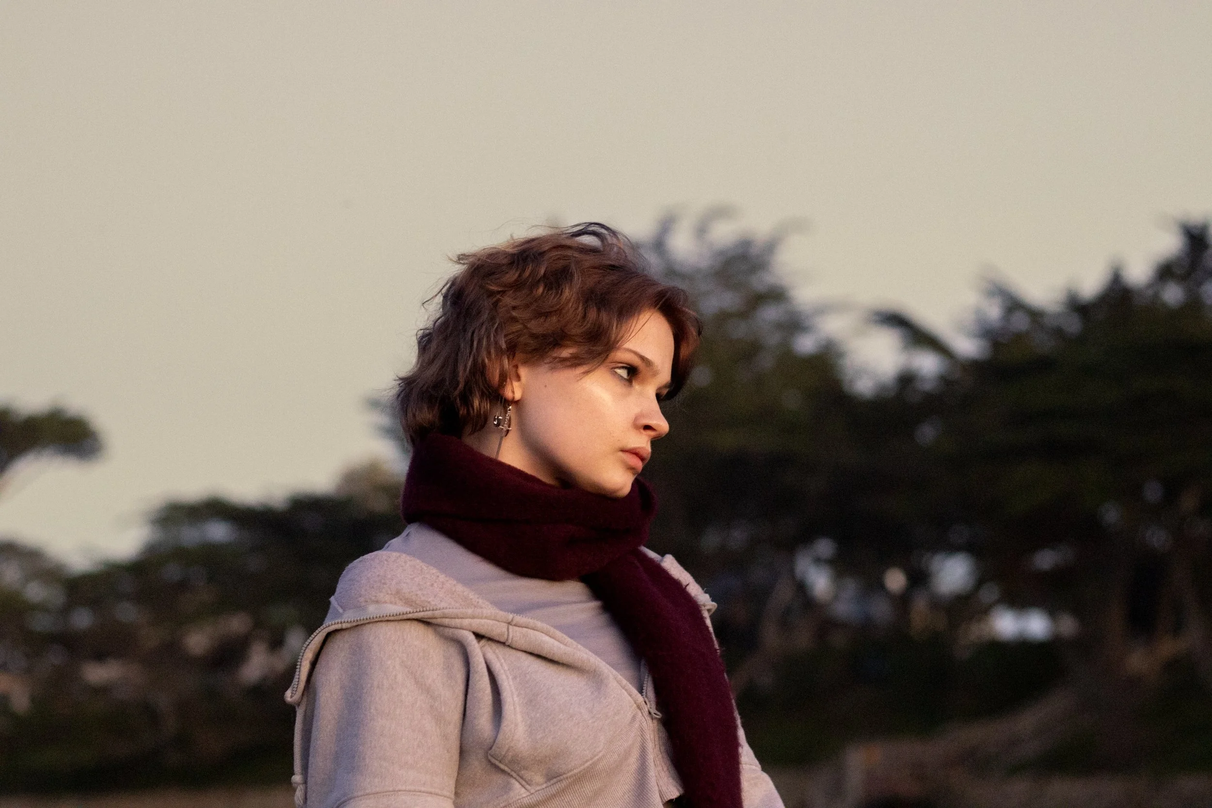 A woman with short, wavy brown hair, wearing earrings, a beige jacket, and a dark red scarf, standing outdoors with a blurred background of trees during dusk or dawn.