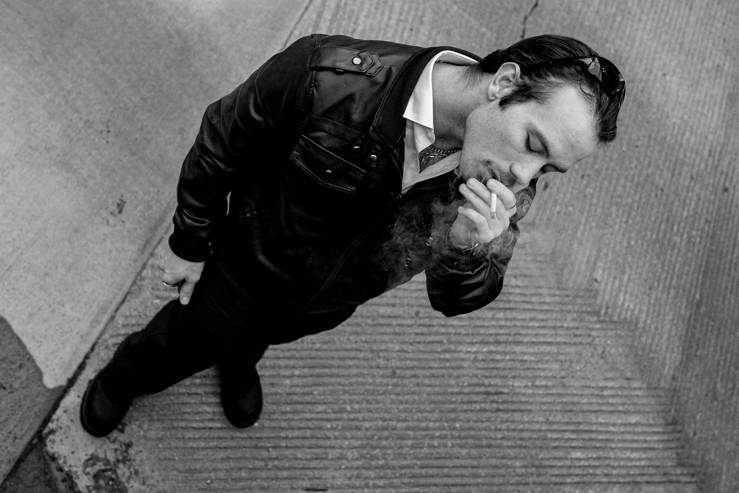 A man standing on the sidewalk, smoking a cigarette, looking down, wearing a leather jacket and sunglasses, seen from an overhead angle.