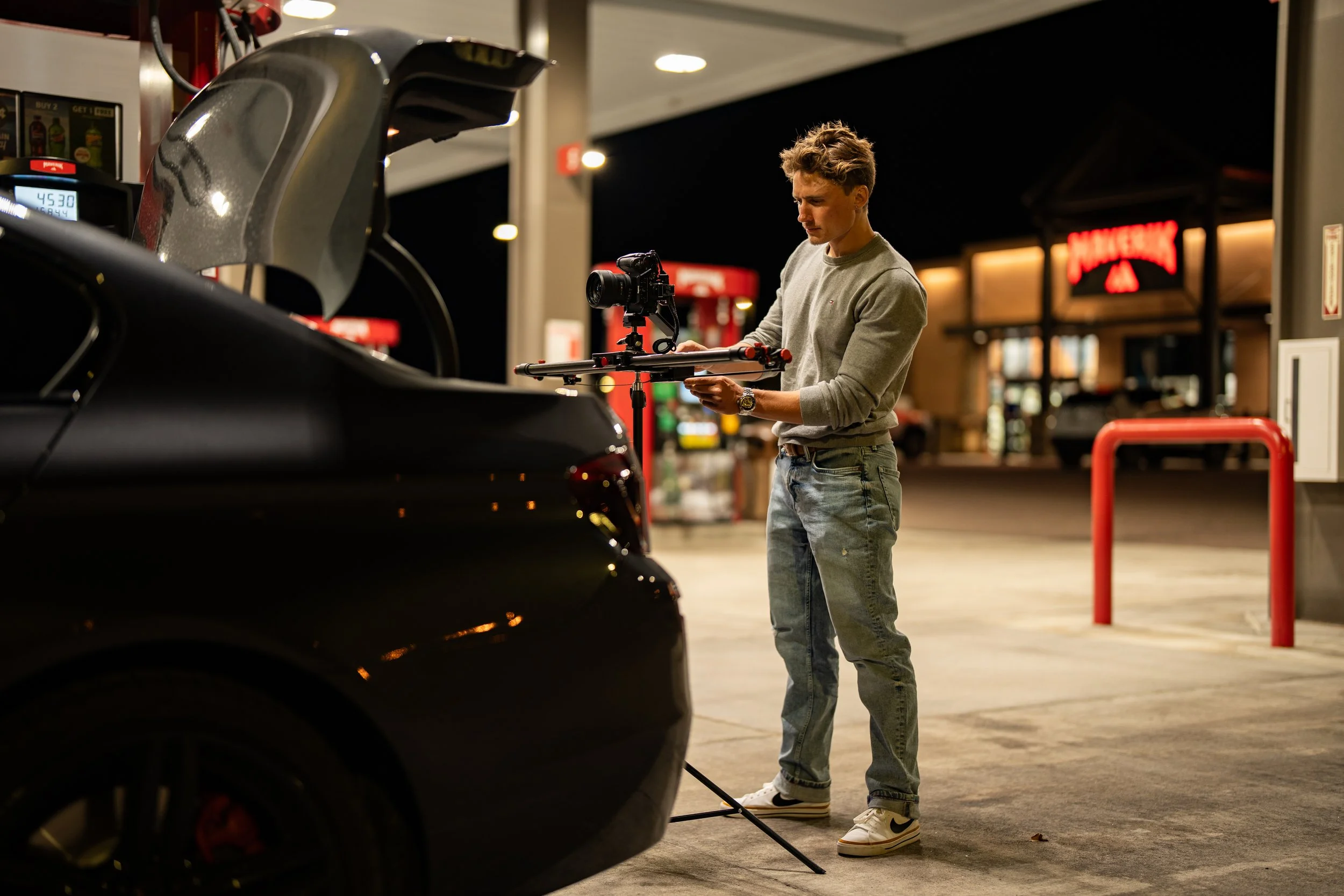 A young man at a gas station at night using a camera mounted on a stabilization rig, next to a black sports car with the gas pump nozzle inserted into the car's gas tank, and a convenience store in the background.