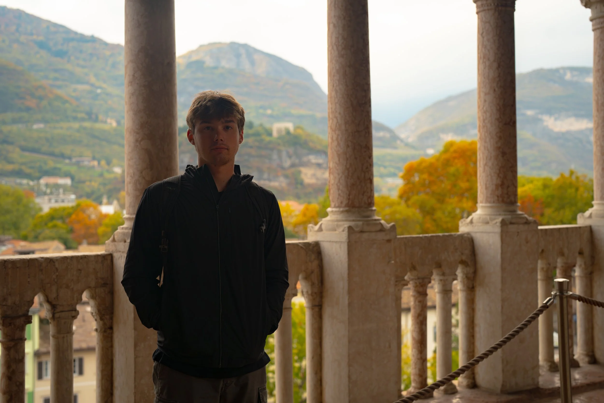 Young man in a black jacket standing on a balcony with stone columns, overlooking a landscape with colorful trees and mountains in the background.
