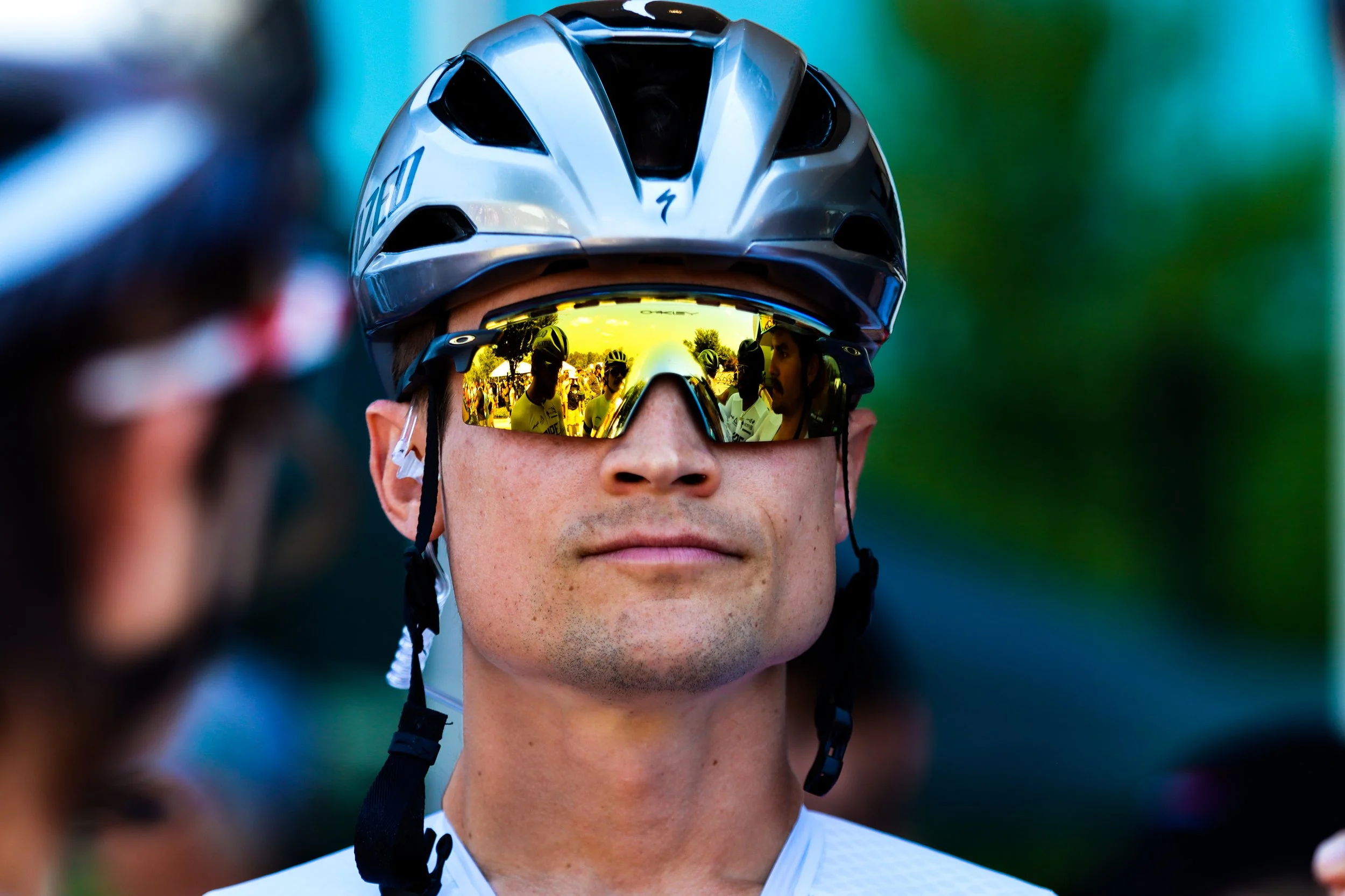 Close-up of a male cyclist wearing a silver helmet and mirrored sports sunglasses, with a reflection of other cyclists and trees in the sunglasses.