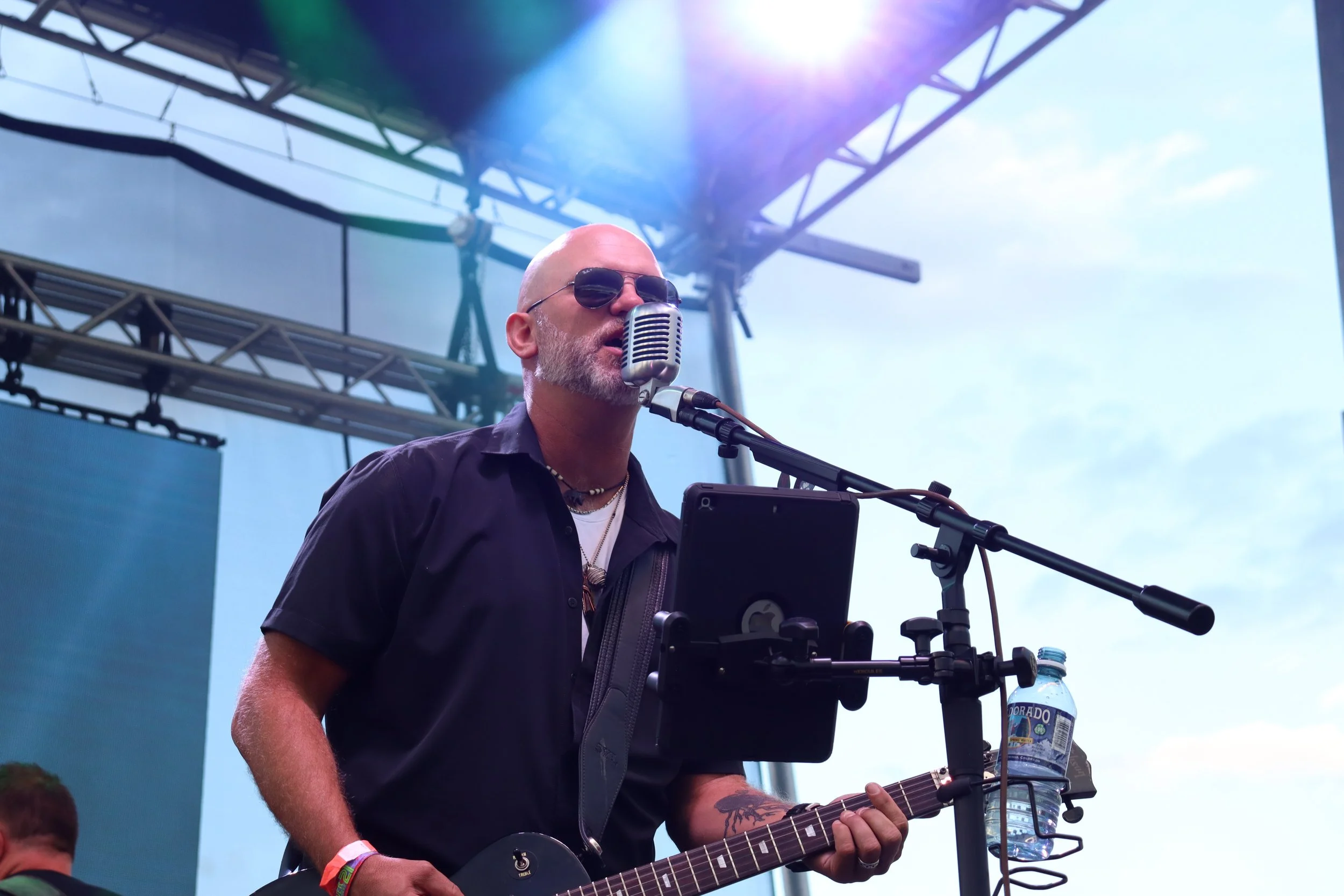 A man with a shaved head and sunglasses singing into a microphone at an outdoor concert, playing an electric guitar, with a bottle of water mounted on a stand next to him and a cloudy sky in the background.