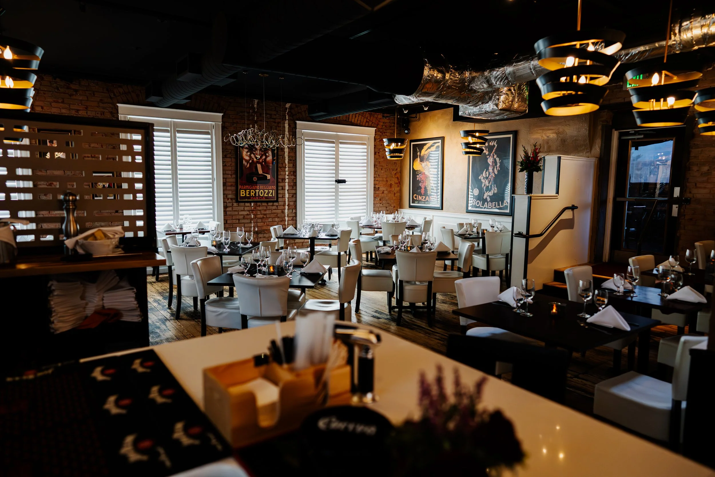 Interior of a restaurant with white chairs and black tables set with wine glasses, napkins, and silverware, brick walls, framed posters, and modern lighting fixtures.