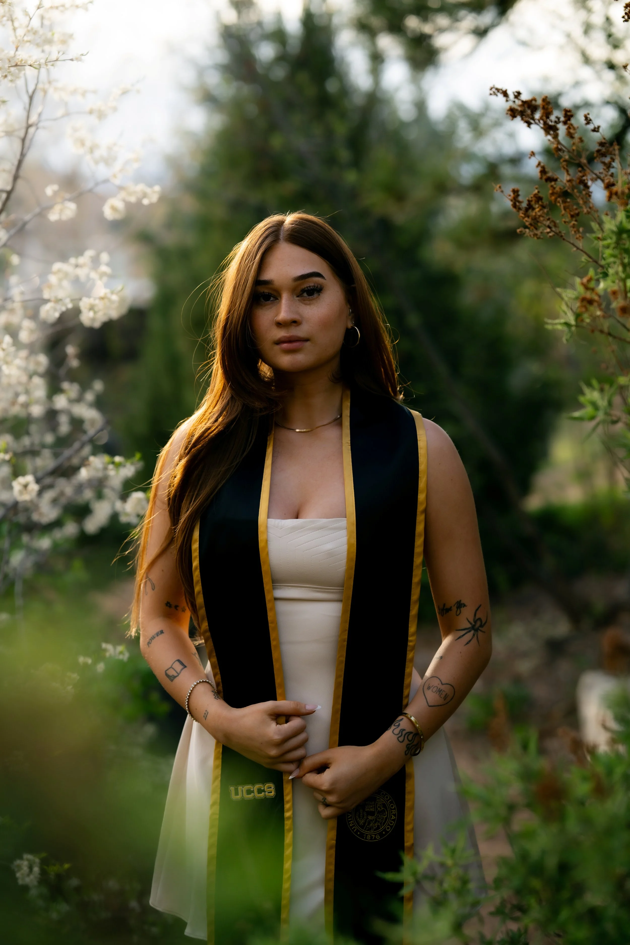 A young woman in a graduation gown standing outdoors amid blooming trees and greenery.