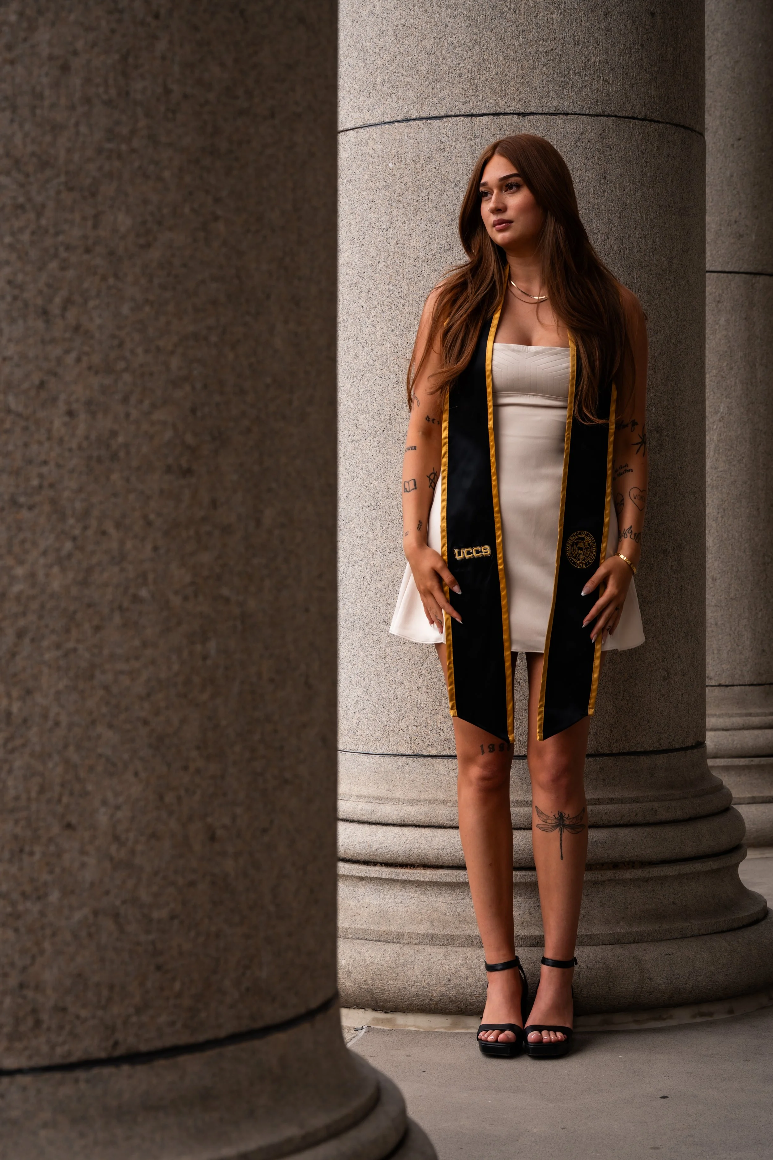 A woman in a white dress and black heels standing between large stone columns, wearing a graduation stole.