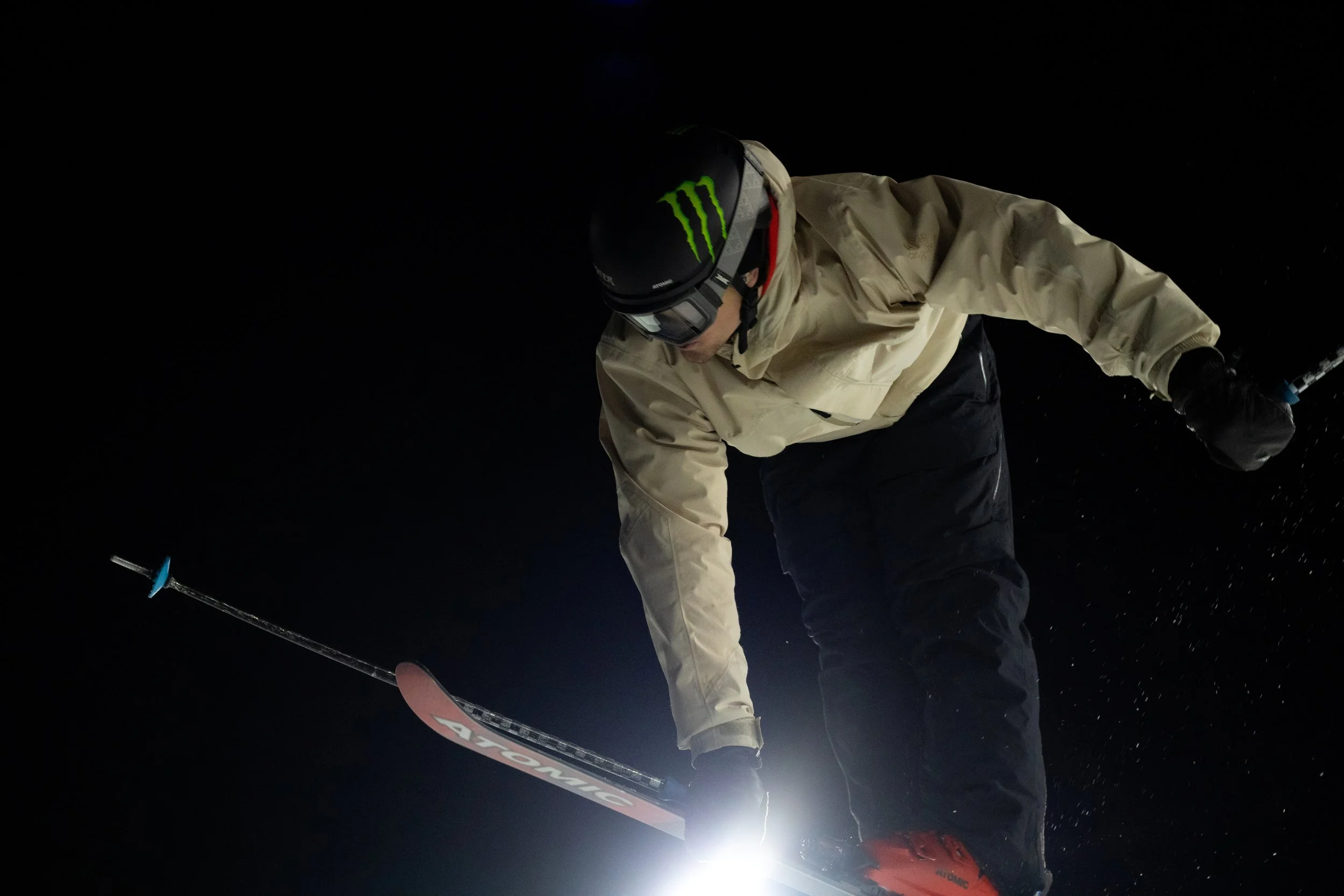 A skier at night on a slope, wearing a beige jacket, black pants, a helmet with a Monster energy logo, goggles, and red ski boots, holding ski poles.