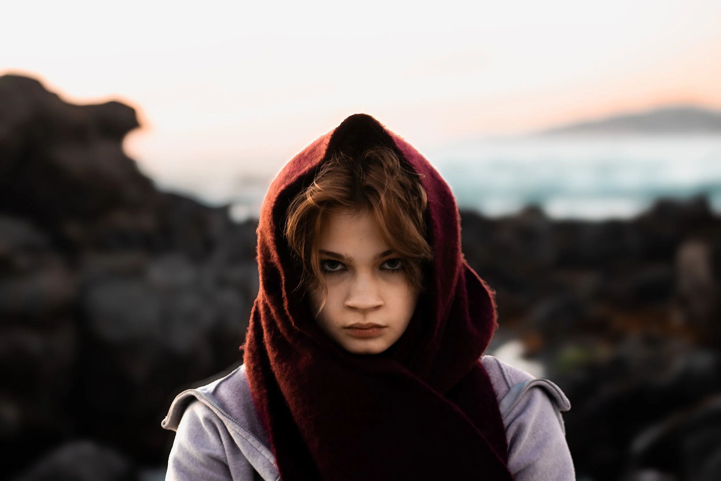 A young woman with short, wavy brown hair and intense eyes stands outdoors during sunset, wearing a deep red hooded scarf and a light gray jacket, with rocky terrain and the ocean blurred in the background.