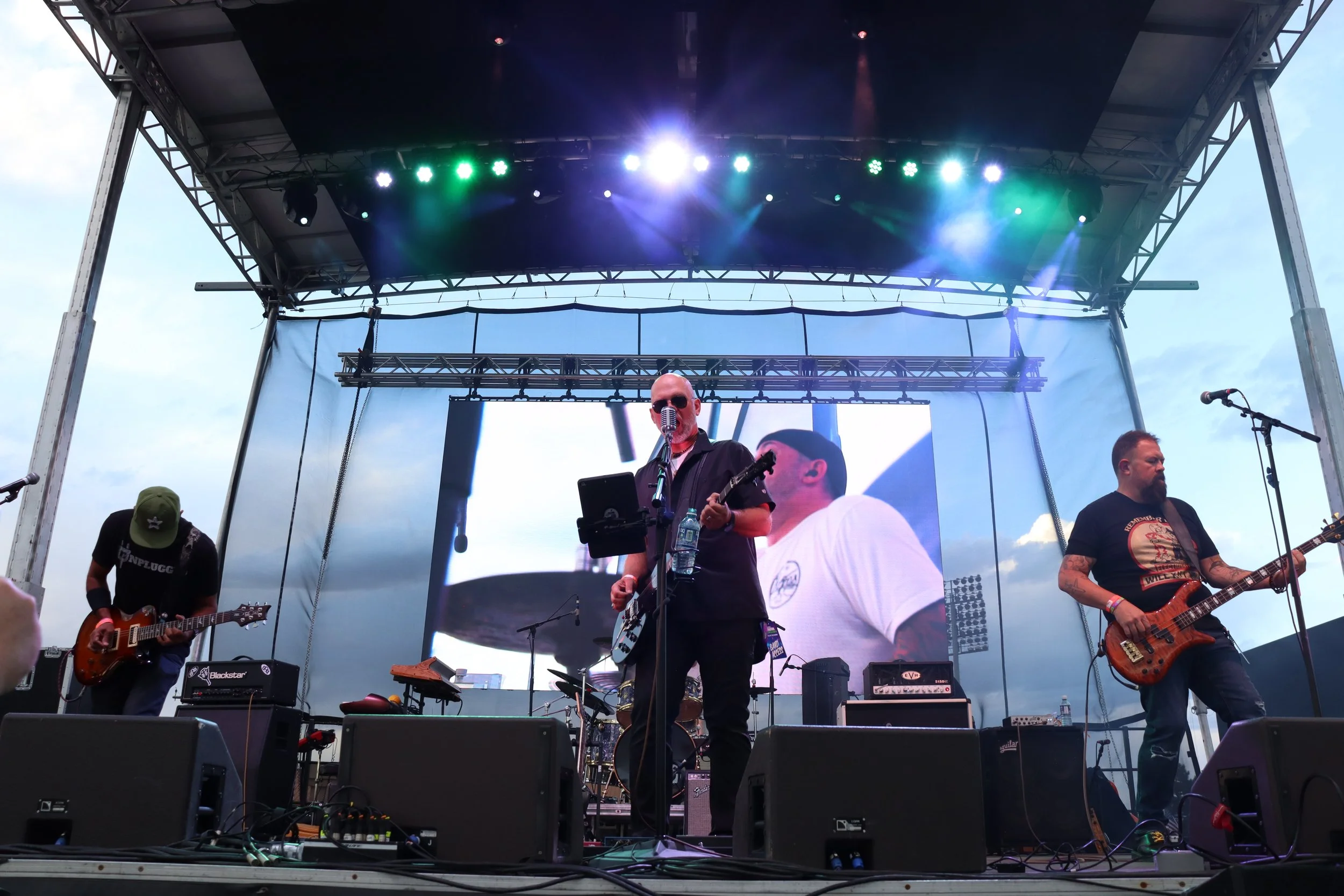 A band performing live on an outdoor stage under colorful lights with a large screen behind them.