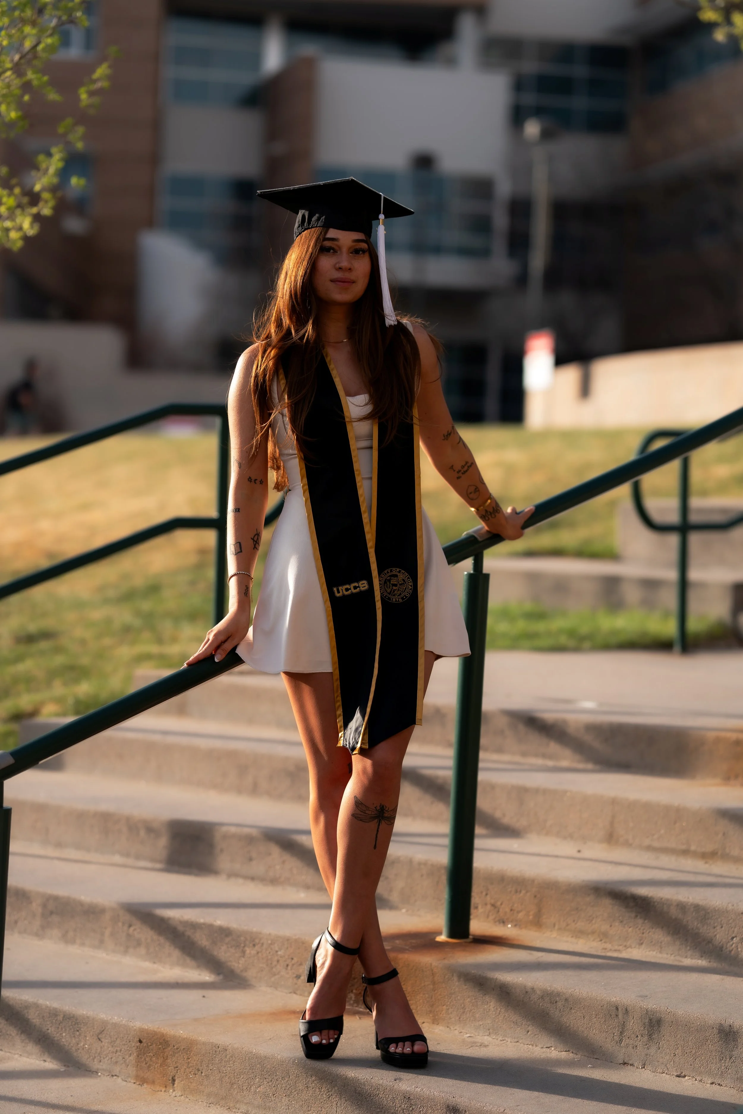 A young woman in a white dress and high heels wearing a black graduation cap and gown stole, standing on outdoor stairs during sunset.