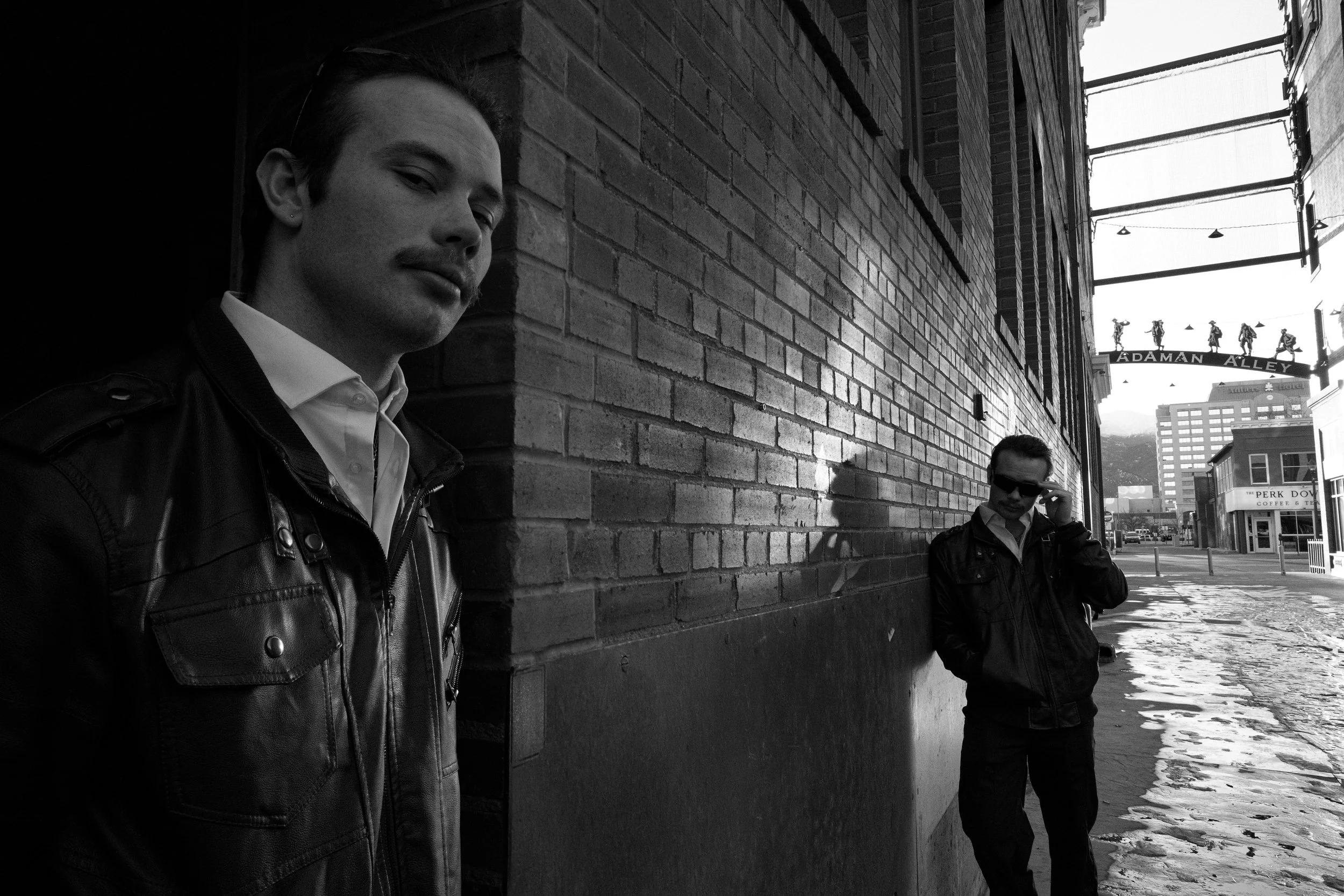 Two men in leather jackets on a city street, one standing close to the camera with a serious expression and the other adjusting sunglasses, with a brick building and street signs in the background.