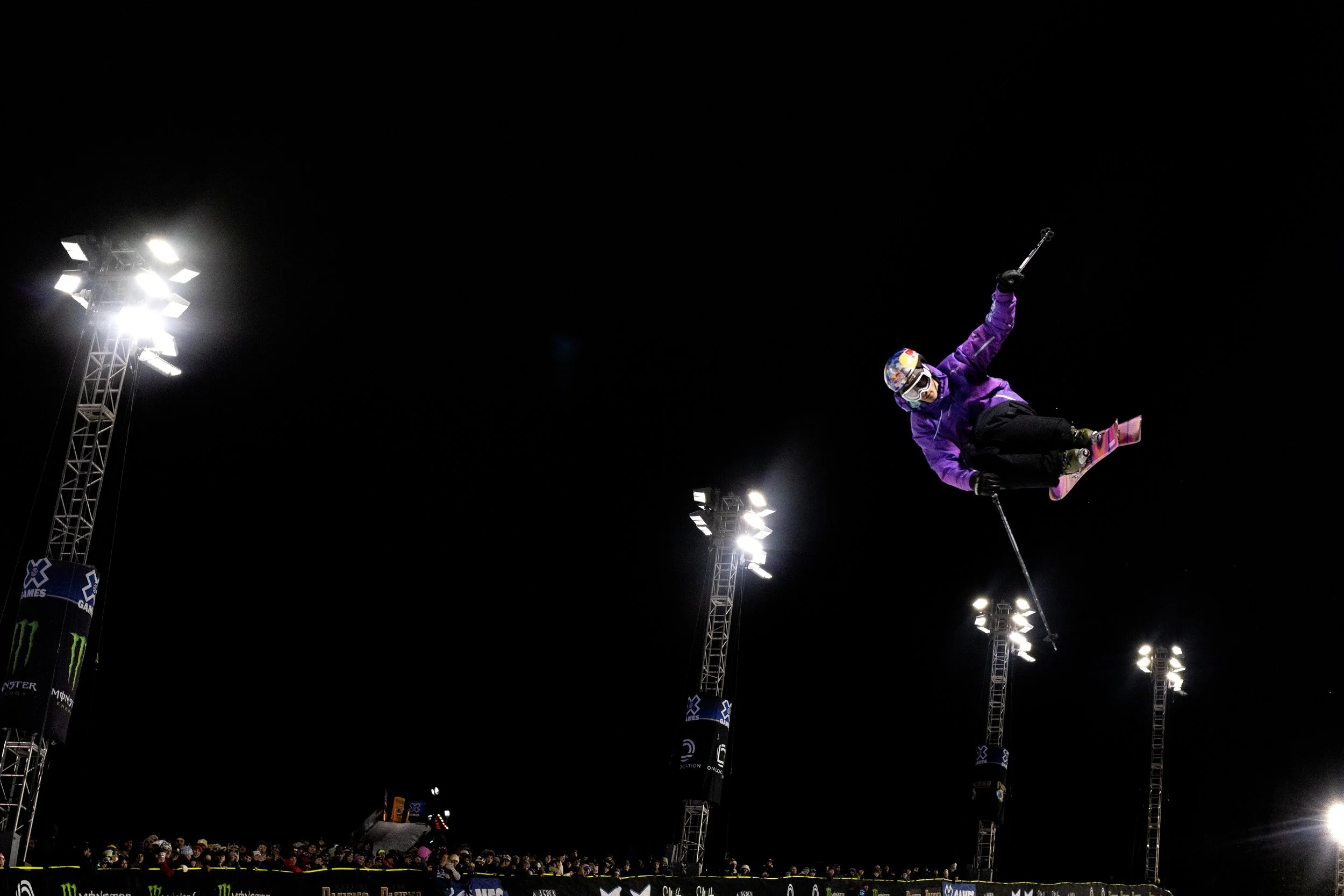 A skier in purple gear mid-air performing a trick at a nighttime event with bright lights and an audience in the background.