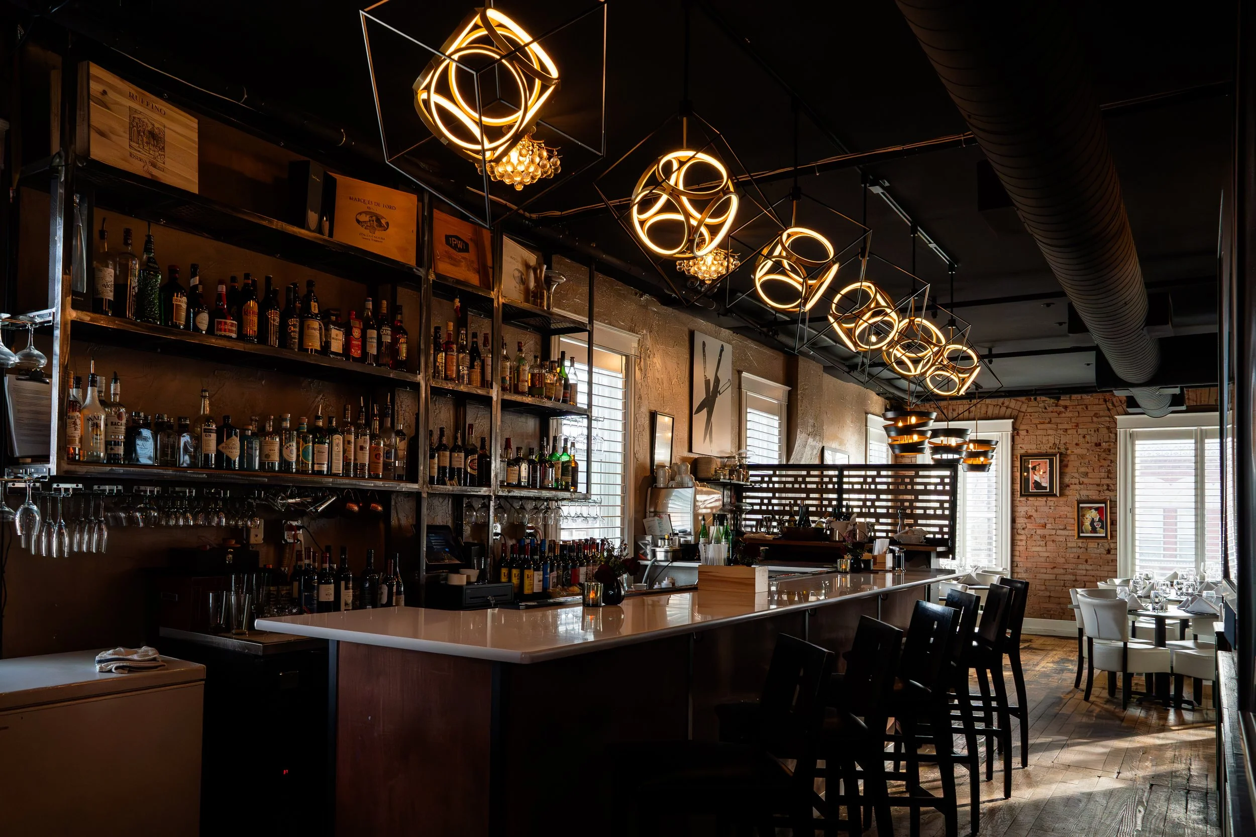 Interior of a modern restaurant or bar with a well-stocked bar, contemporary lighting fixtures, and seated dining area with white chairs near windows.