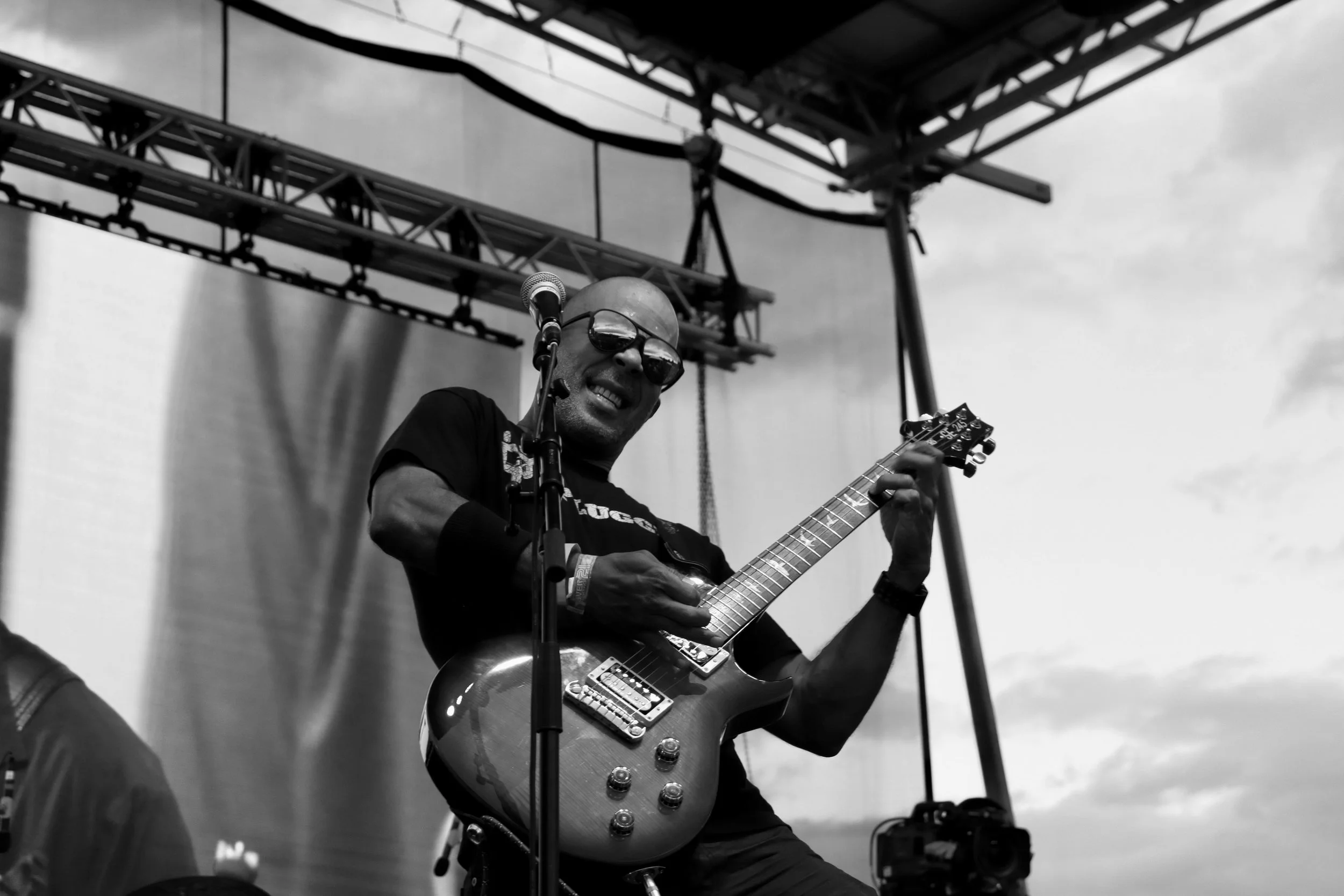 A man wearing sunglasses and a black T-shirt playing an electric guitar on an outdoor stage during a performance.