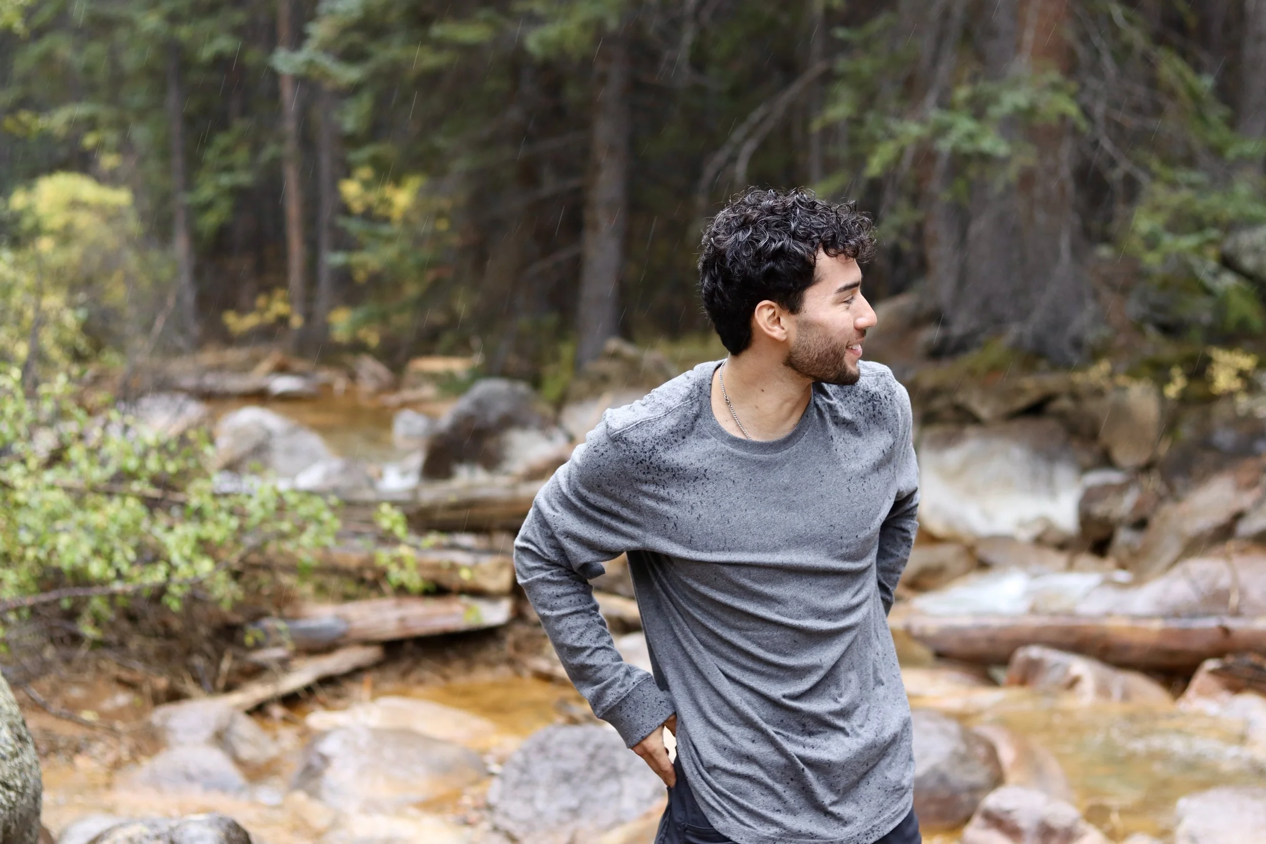 A man standing outdoors near a creek in a forest, with wet hair and shirt, smiling and looking to the side.