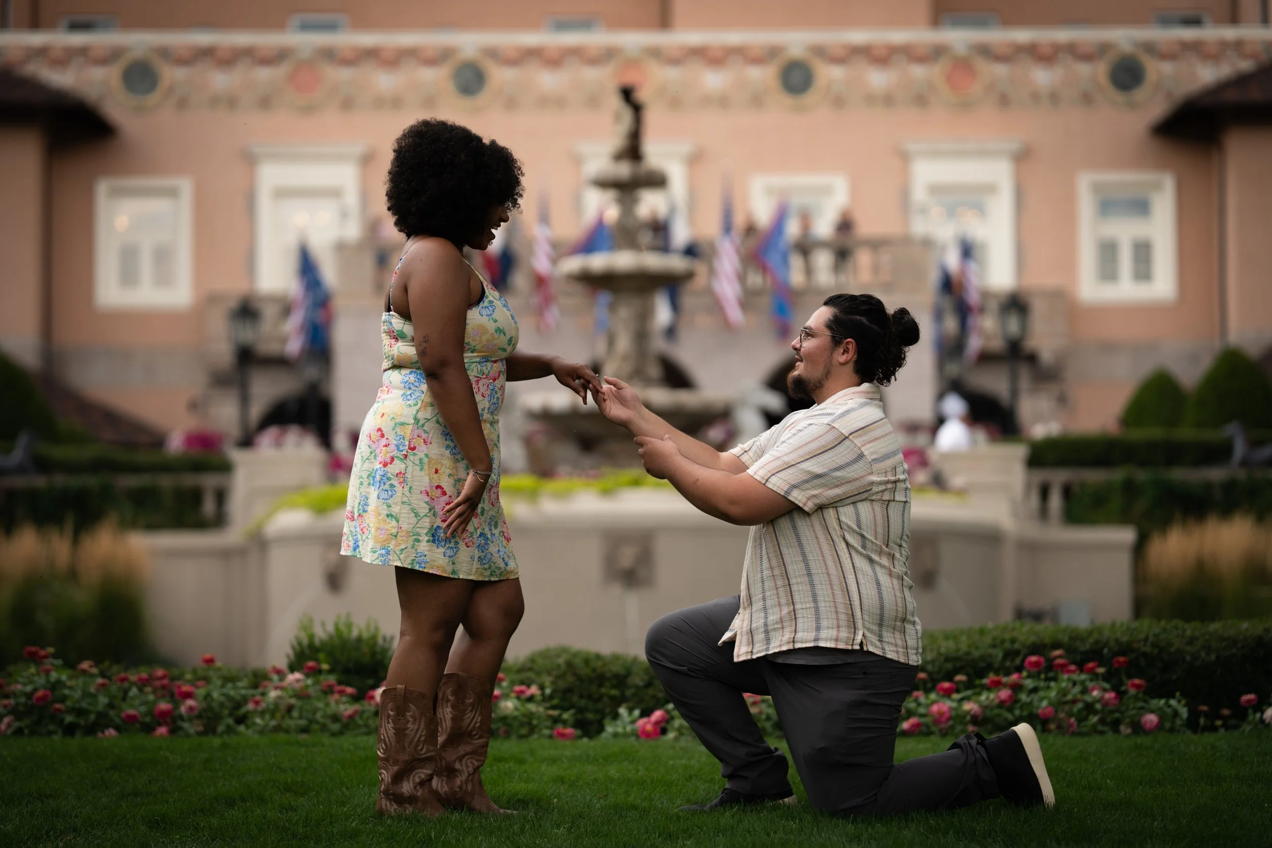 A man is kneeling on one knee proposing to a woman in a garden in front of a fountain and a pink building with American flags.