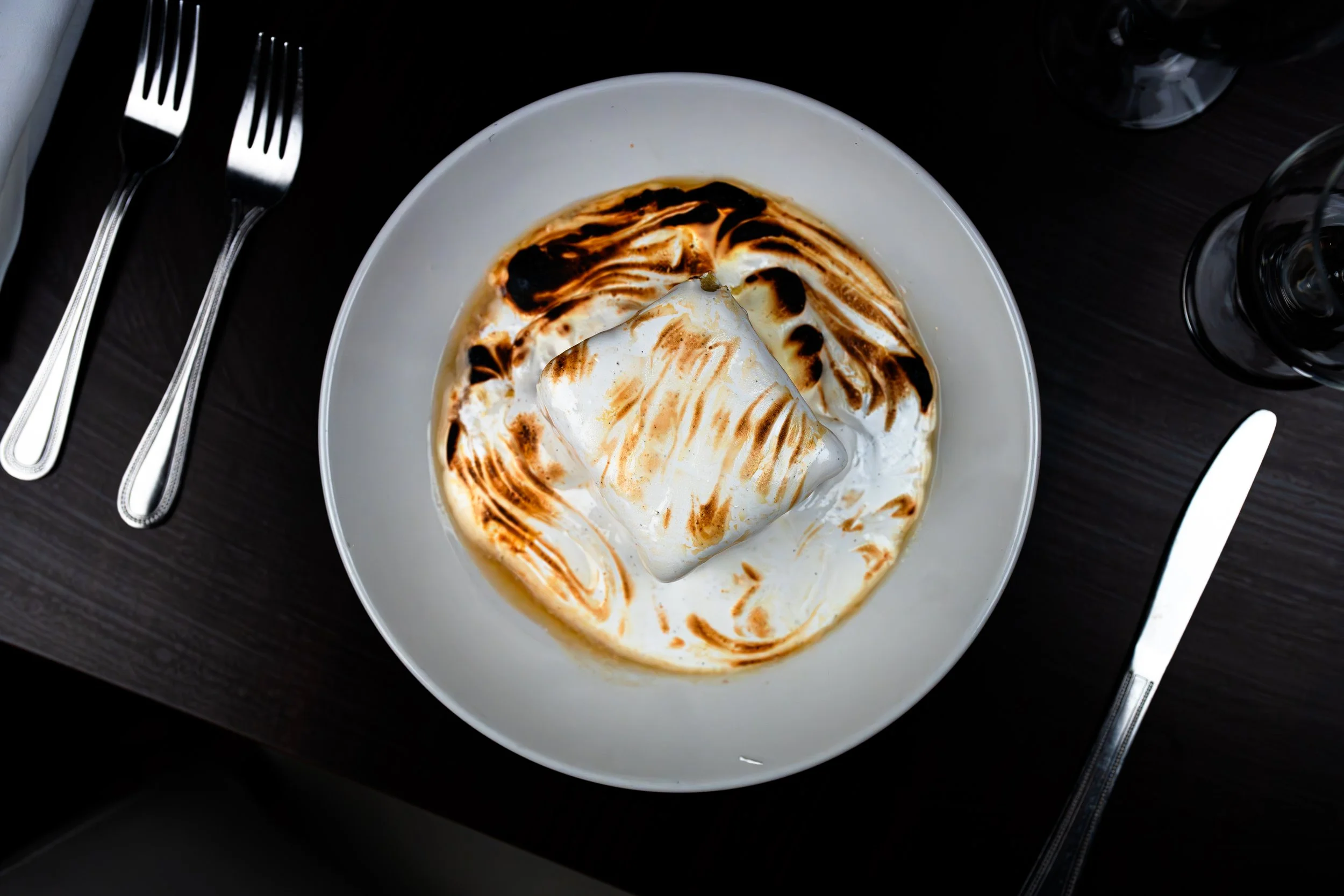 A bowl of dessert topped with toasted marshmallow and chocolate sauce on a dark table, with a fork on the left and a knife on the right.