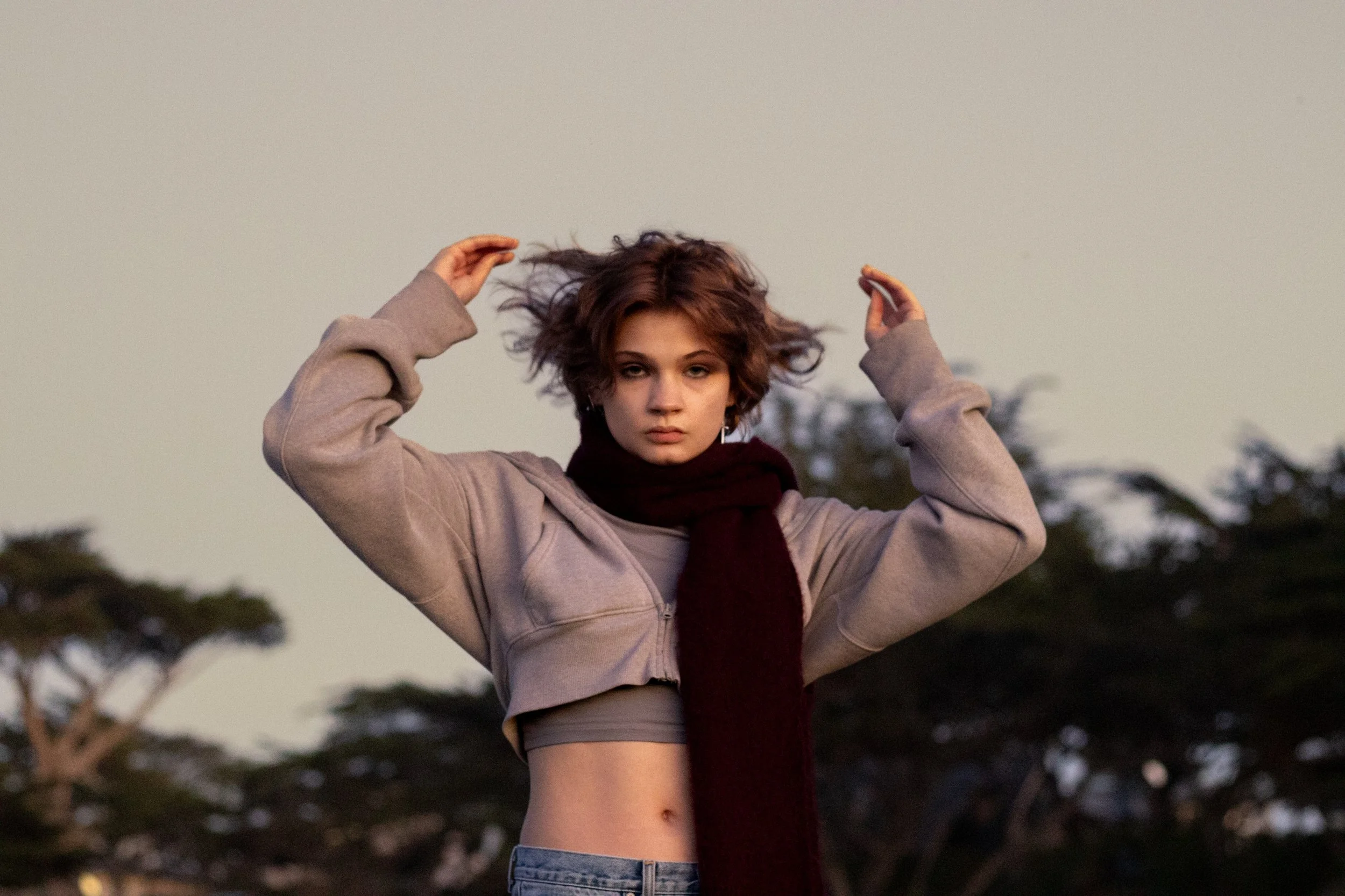 A young woman with short, curly brown hair stands outdoors against a background of trees, wearing a beige cropped jacket, a gray top, a dark red scarf, and jeans, with her arms raised and her hands near her head.