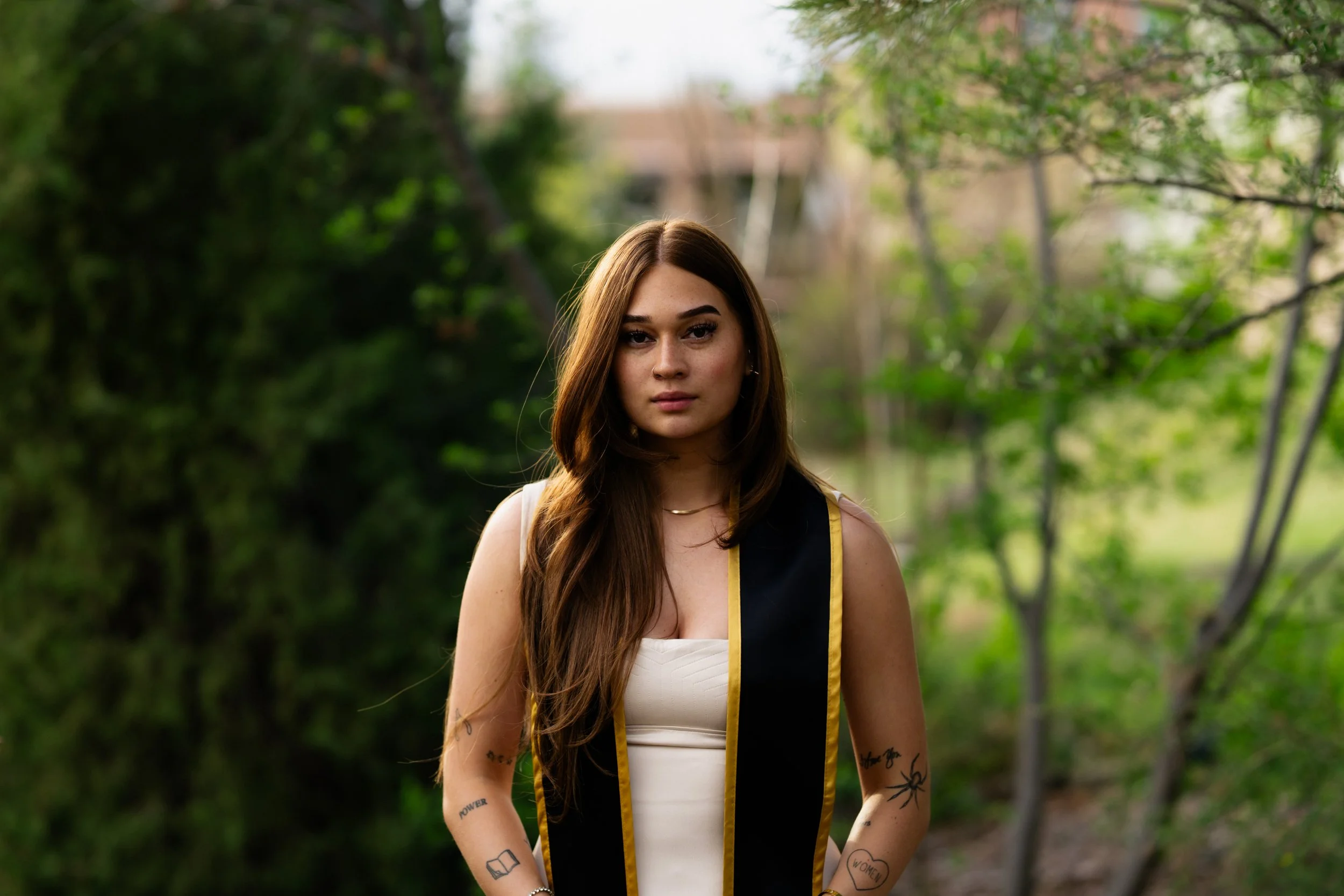 Young woman with long brown hair wearing a graduation stole, standing outdoors in a green, wooded area.