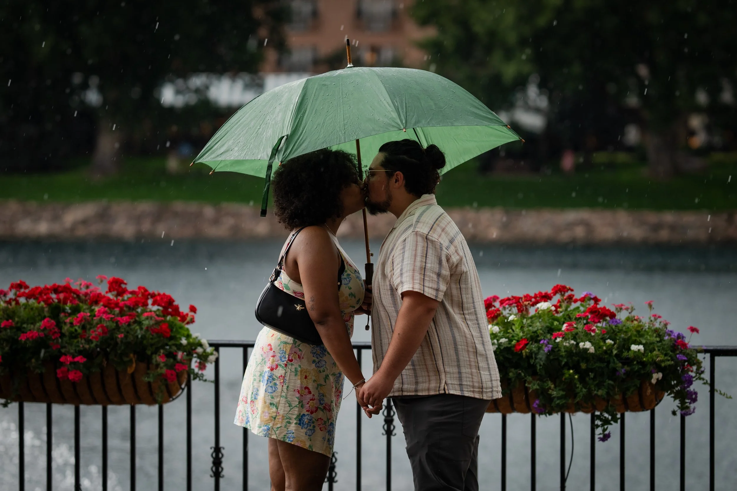 A couple sharing a kiss under a green umbrella by a lake with colorful flowers on a rainy day.