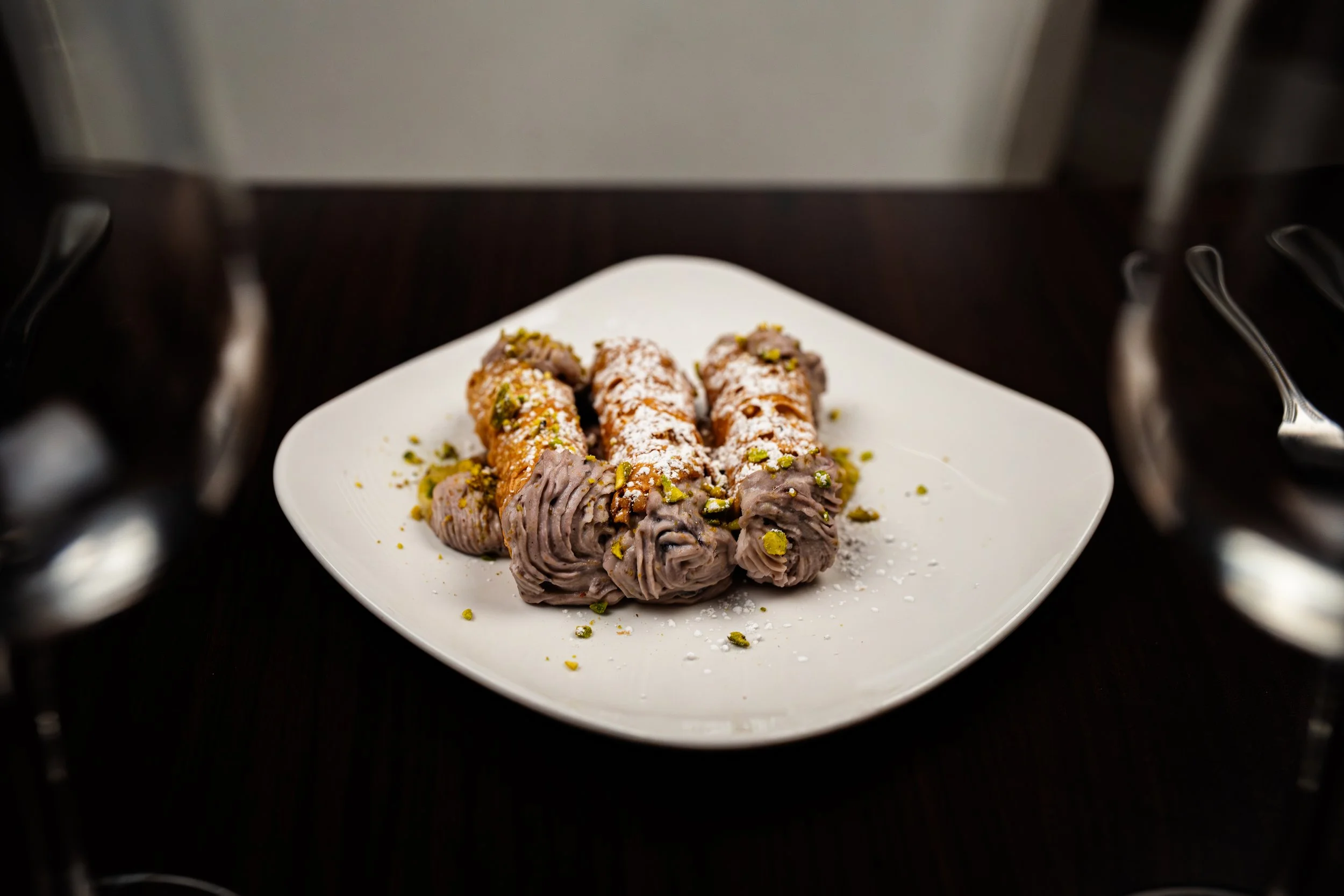Plate of four cannoli with chocolate filling, sprinkled with powdered sugar and chopped pistachios, on a white square plate on a dark table.