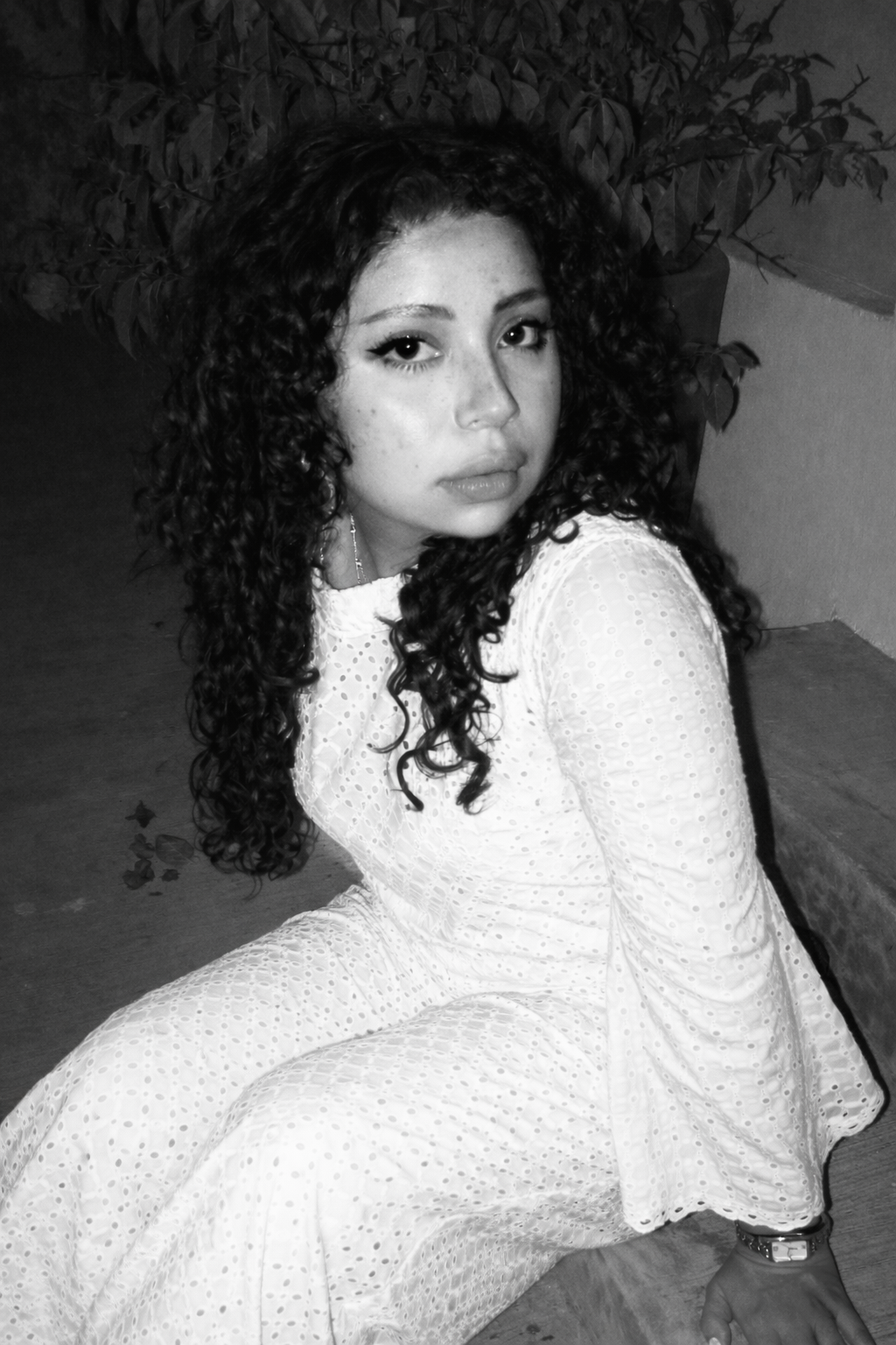 A young woman with curly hair and freckles sitting on the floor, looking at the camera, wearing a white dress with eyelet pattern, with a potted plant behind her.
