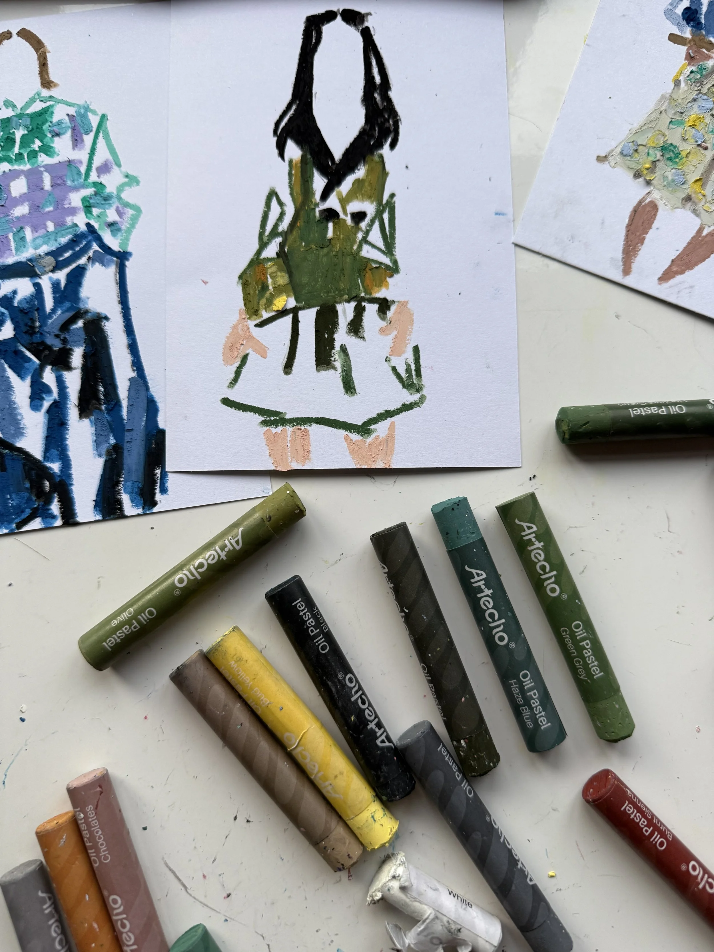 Children's artwork featuring a girl in a green dress with a black hairstyle, painted with oil pastels, surrounded by other colorful drawings and pastels on a white table.