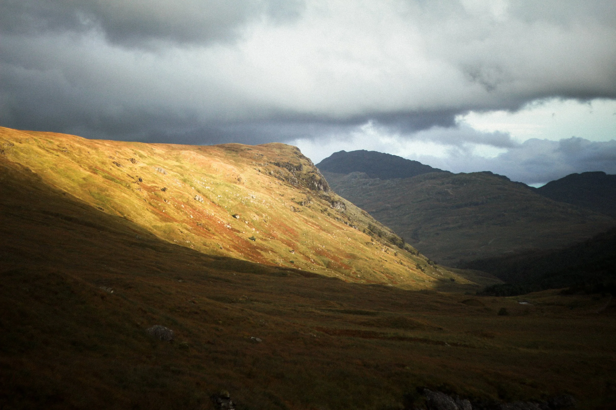 Hilly landscape with green and brown grass, dark clouds overhead, and distant mountains.