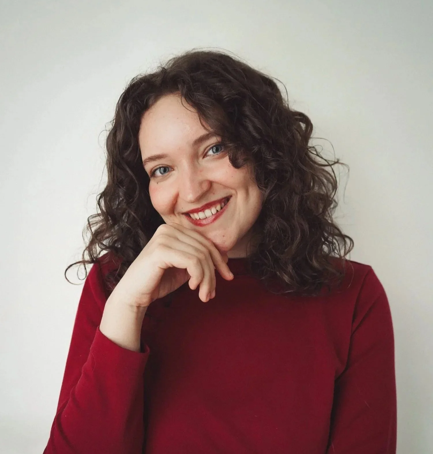 A woman with curly dark hair and blue eyes, wearing a red top, smiling and resting her chin on her hand against a plain white background.