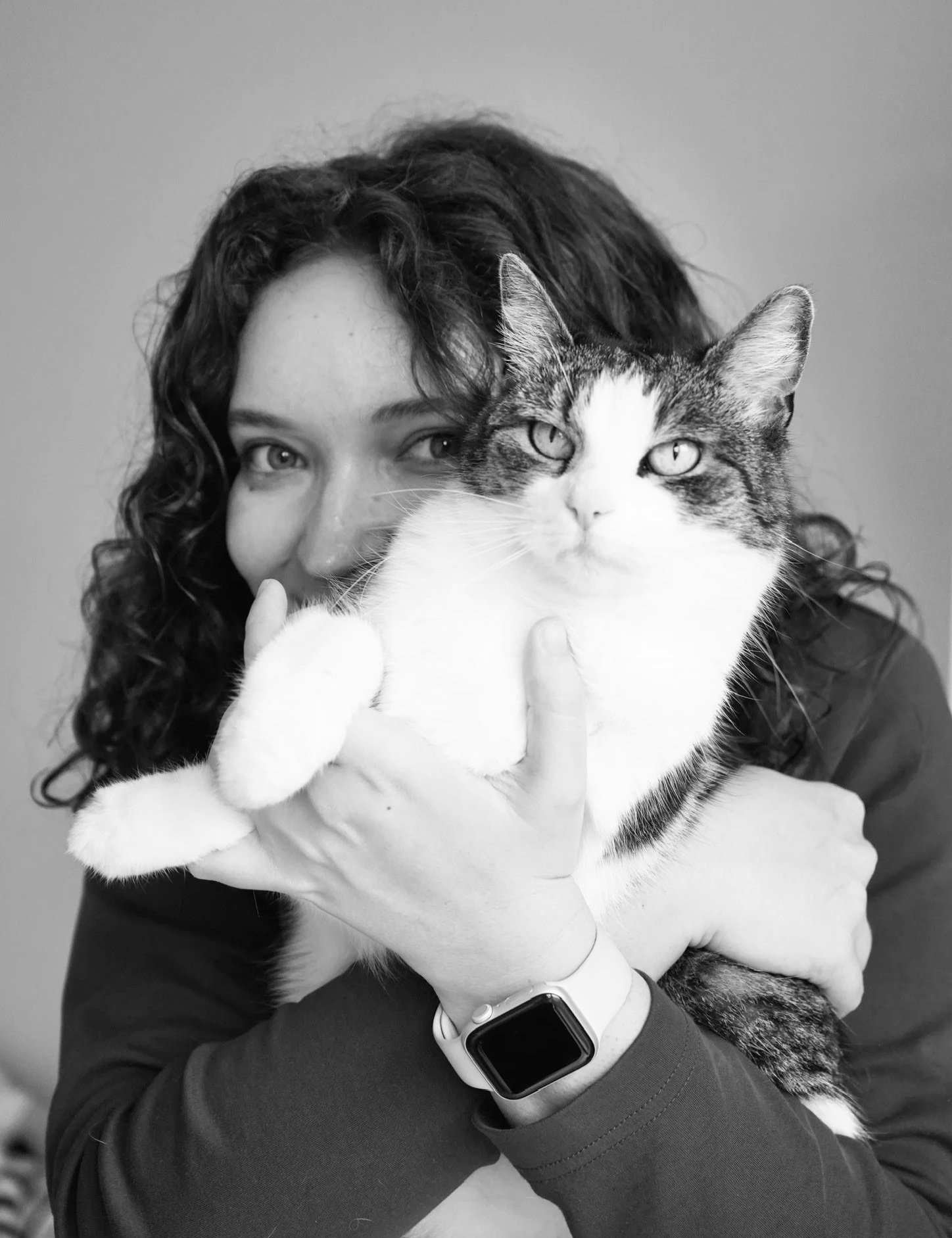 A woman with curly hair holding a tabby and white cat close to her face, both looking at the camera.