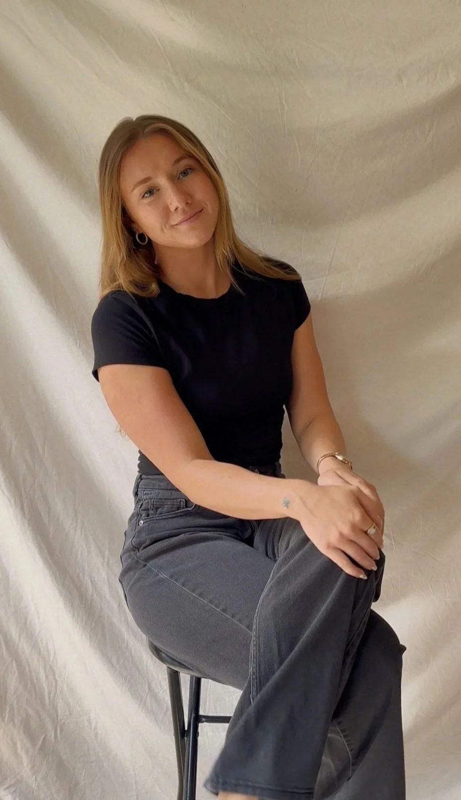 A young woman with shoulder-length blonde hair and blue eyes, dressed in a black t-shirt and gray jeans, sitting on a black stool against a beige textured background.