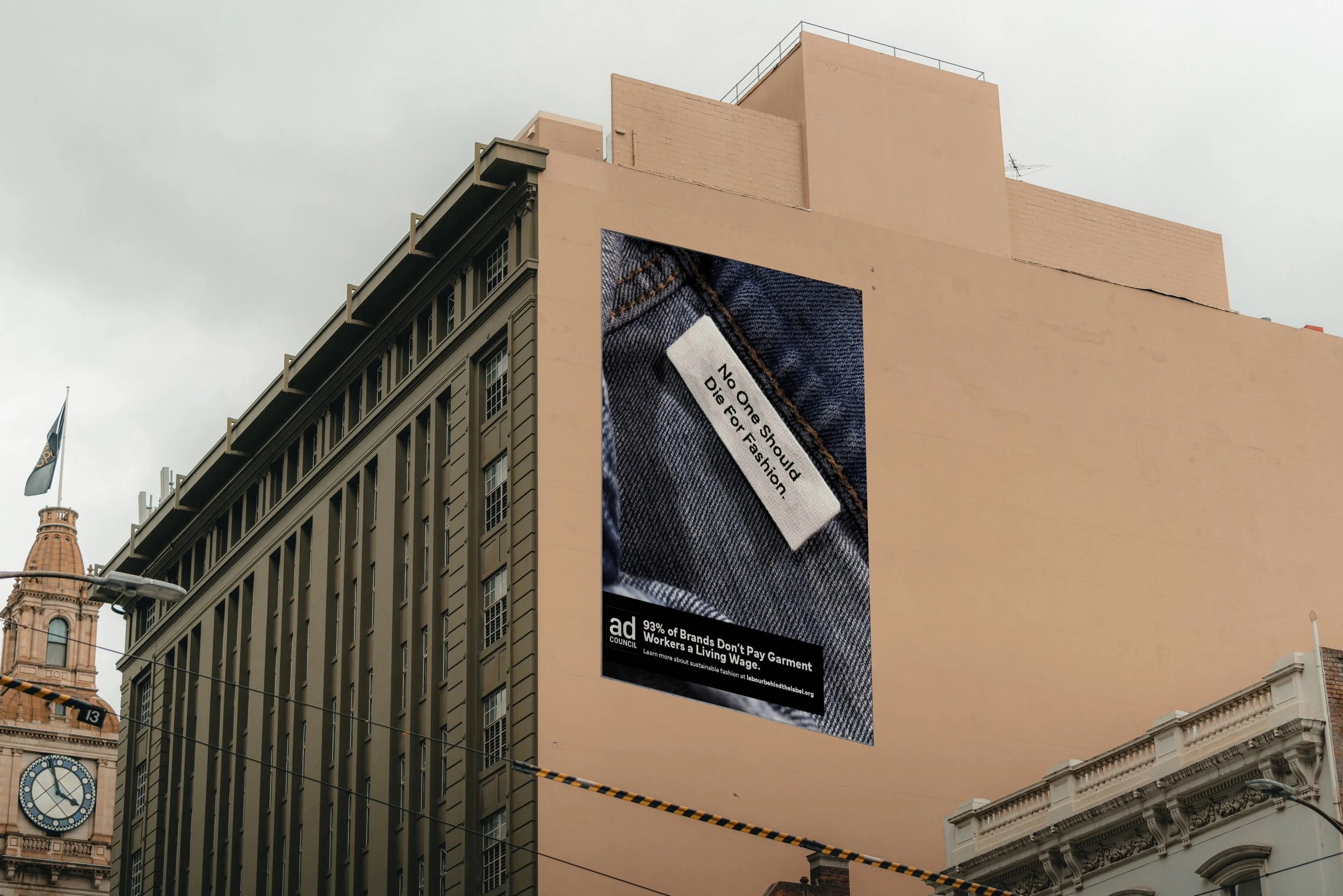 A city street scene with a large outdoor billboard displaying a close-up of denim fabric and a clothing label that reads "No One Should Dig For Fashion." The surrounding architecture includes historic and modern buildings, with a clock showing 12:12 and a flag on a flagpole.