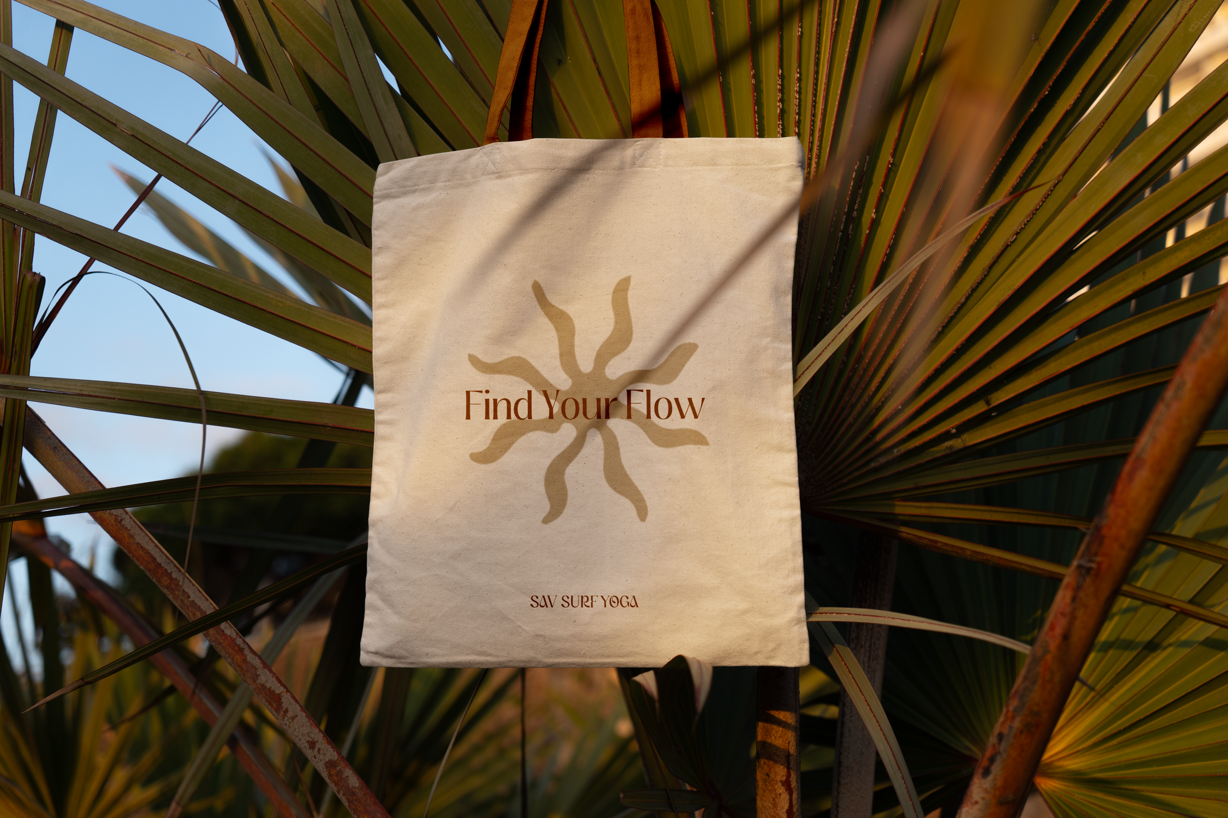 A beige tote bag with the phrase 'Find Your Flow' and a sunburst logo, hanging on a rusty metal fence among green palm leaves in bright daylight.