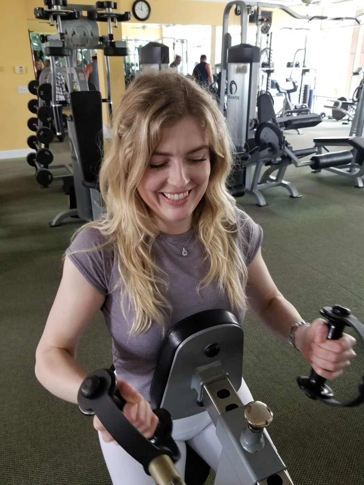 A woman smiling while using a stationary exercise bike in a gym. She has long blonde hair and is wearing a gray shirt. There are various gym equipment and mirrors in the background.