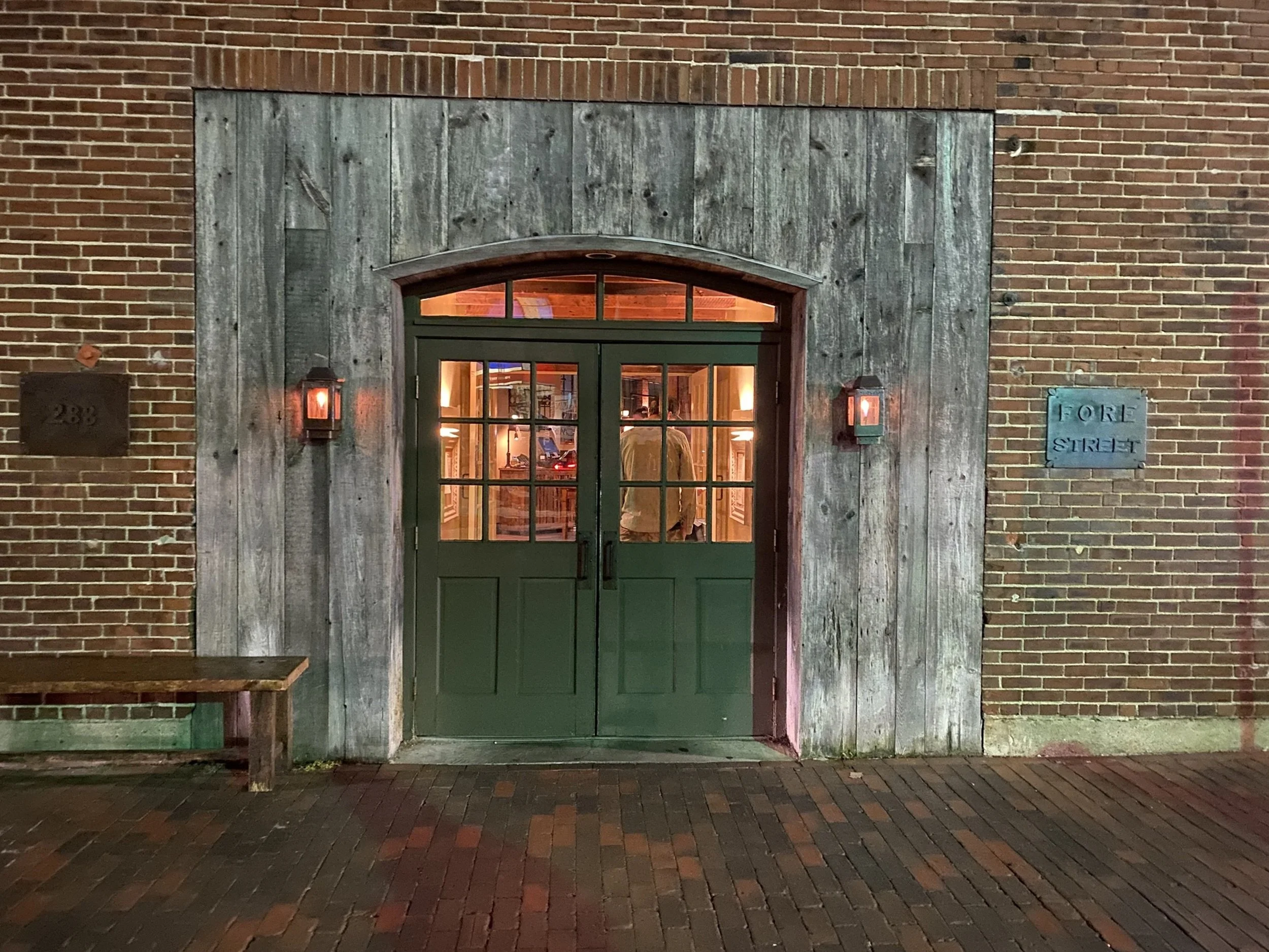 Fore Street - Evening Entrance: Warm light spills through a historic doorway framed by weathered timber and brick, creating an inviting sense of arrival after dusk. Rich in texture and atmosphere, this image is perfectly suited for hotel corridors, l