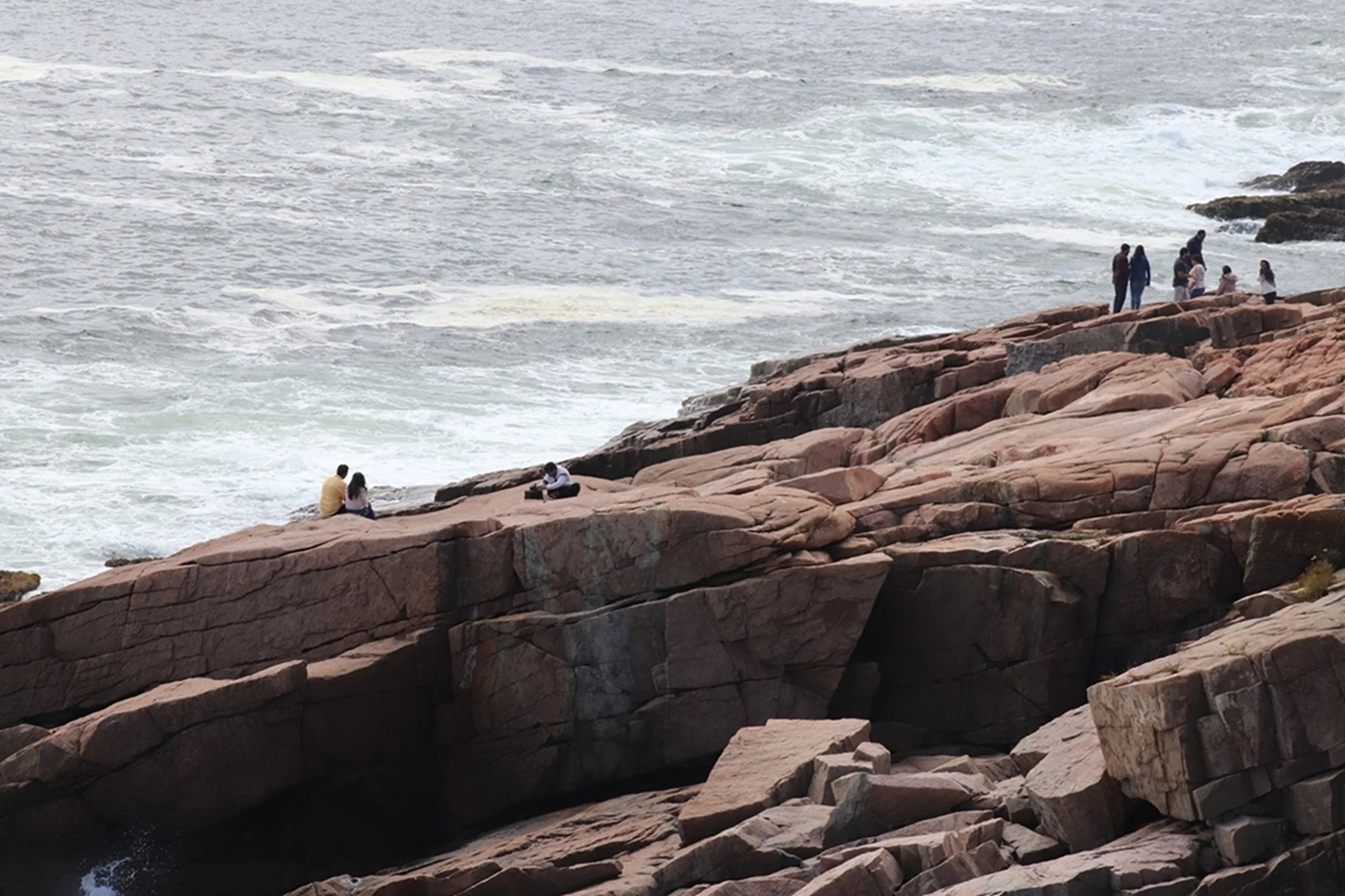 People sitting on rocks by the ocean, some standing and some sitting, with waves crashing onto the rocks.
