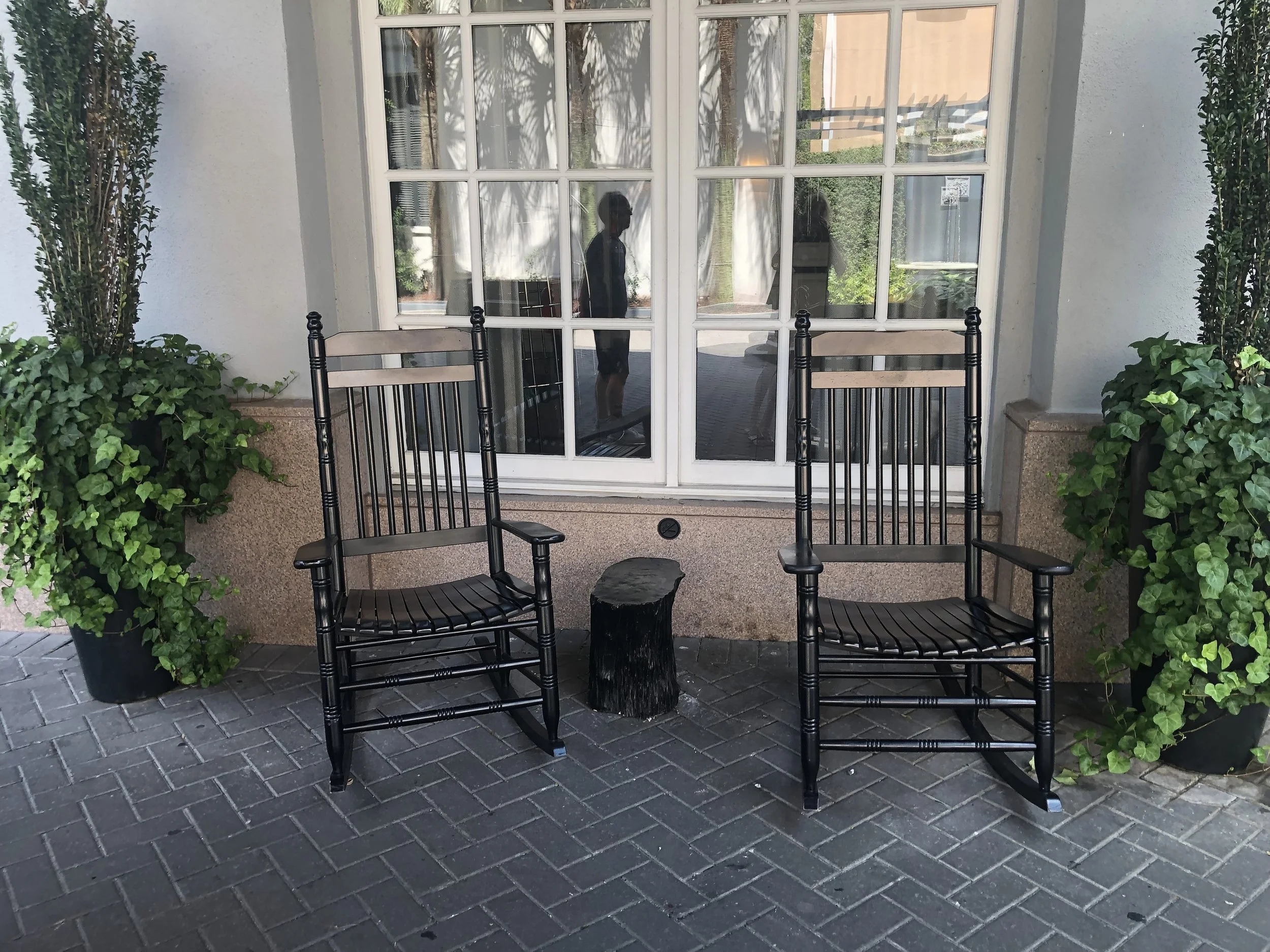 Side by Side

Two black rocking chairs sit symmetrically beneath a paned window, framed by tall greenery. The composition balances structure and softness, suggesting quiet conversation and unhurried moments.

The repetition and restraint create visua