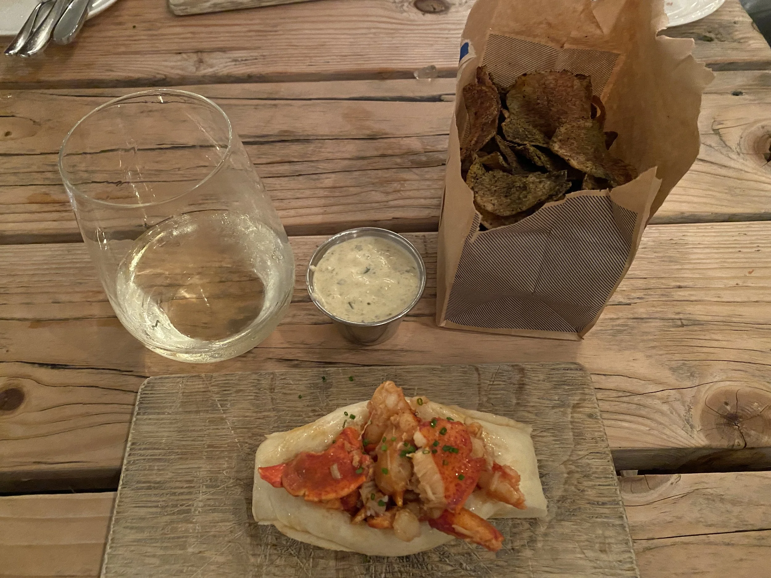 Lobster Roll & White Wine - Coastal Table

A classic lobster roll is presented simply on a wooden board, paired with house-made chips, a glass of white wine, and a small serving of sauce. The composition feels relaxed and approachable, highlighting t