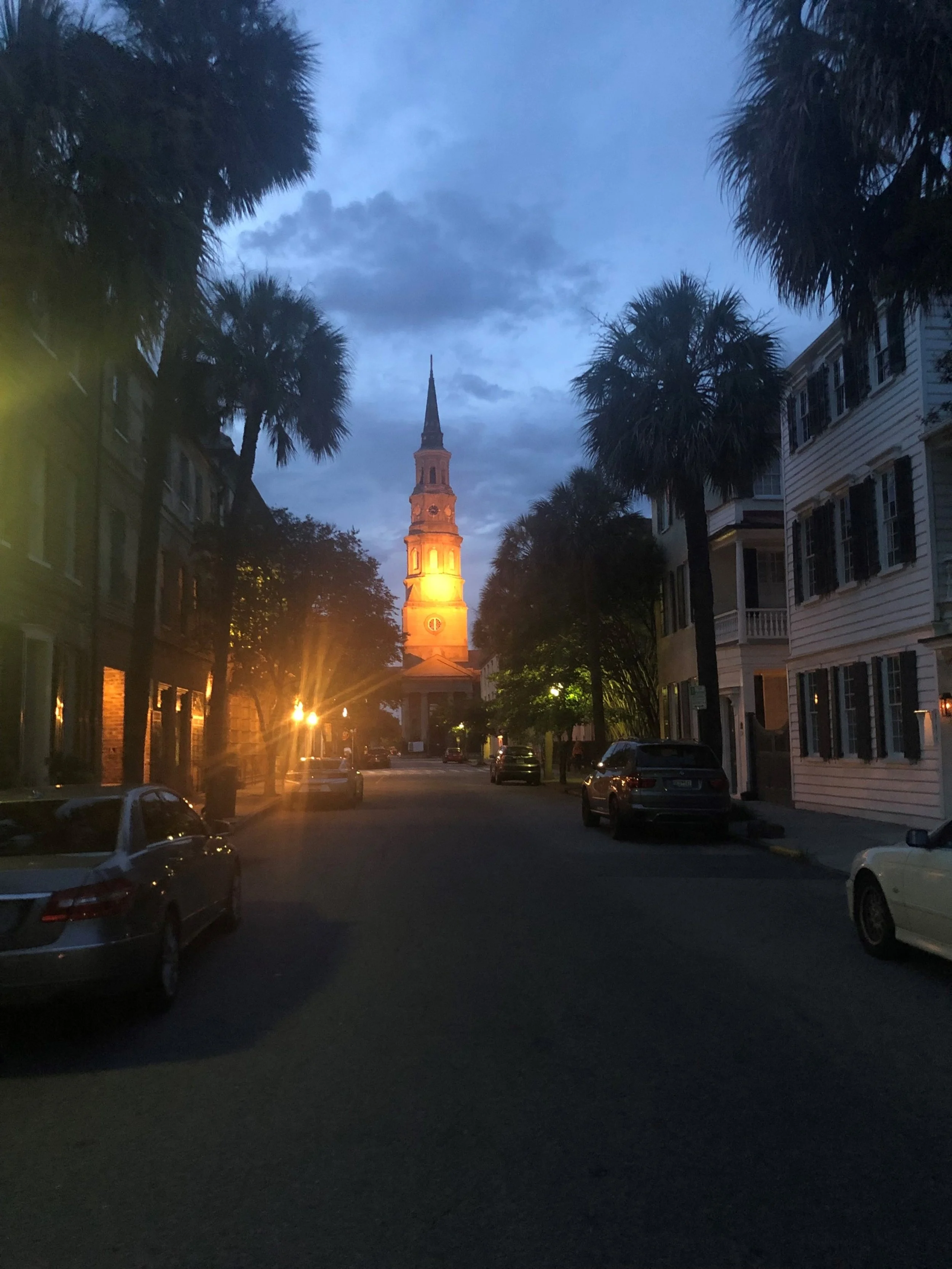 Evening Glow - St. Michael's at Dusk

A softly lit church steeple rises at the end of a quiet street, framed by palms and historic facades as dusk settles in. Warm light from the tower contrasts with the deepening blue sky, creating a cinematic sense