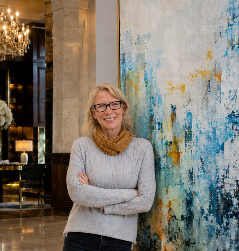A woman with blonde hair and glasses stands with her arms crossed, smiling, beside a colorful abstract painting in an elegant room with a chandelier and warm lighting.