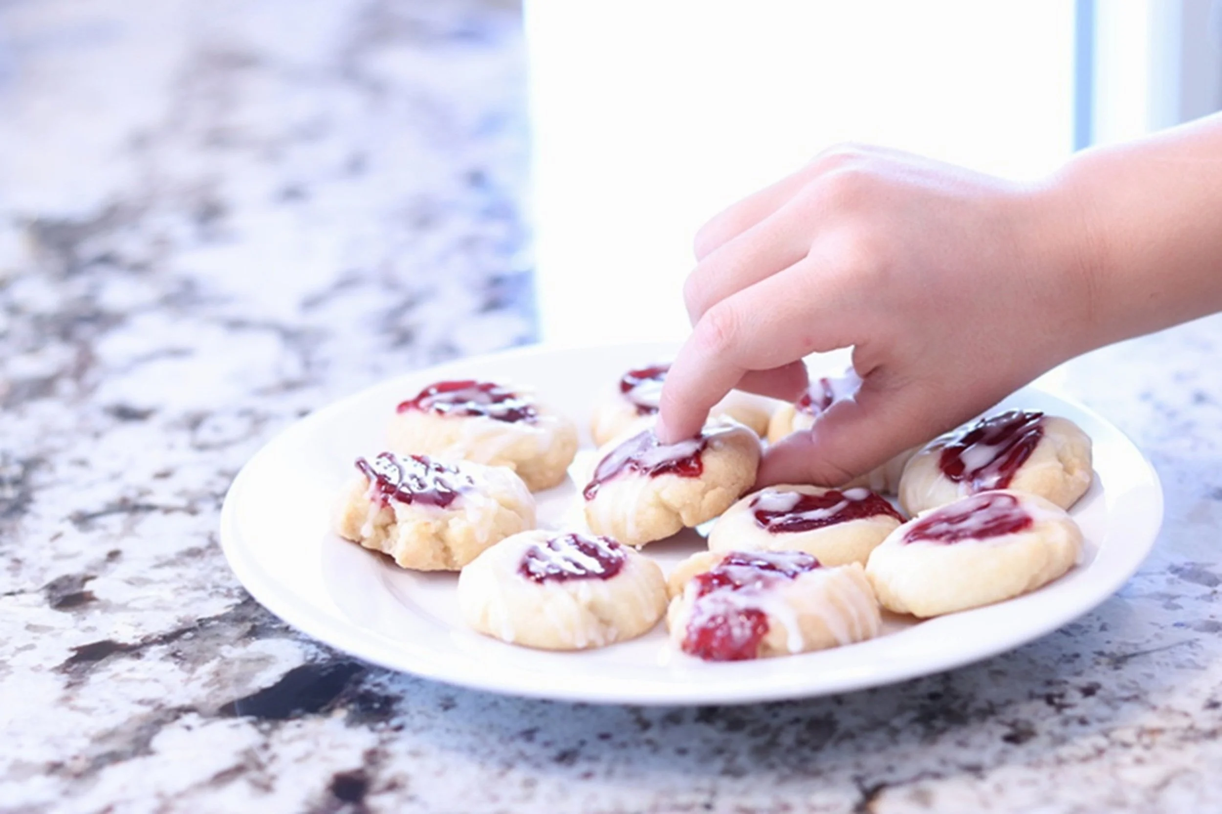 Just One More: A hand reaches for a freshly baked raspberry thumbprint cookie, capturing a spontaneous moment of indulgence and shared enjoyment. The image brings warmth, familiarity, and a welcoming sense of home to cafés, bakeries, breakfast venues