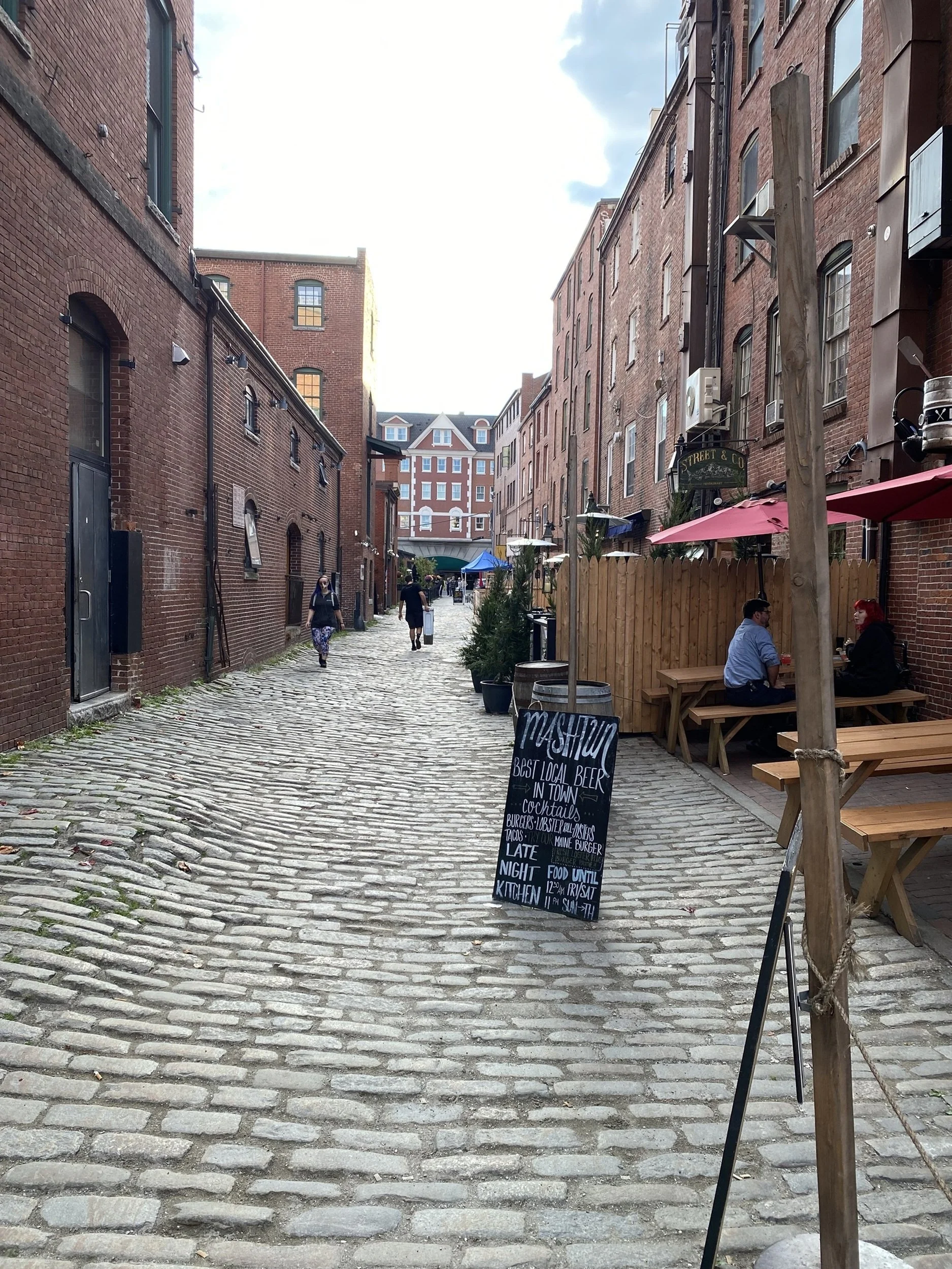 Side Street Social: A cobblestone lane lined with brick buildings, outdoor dining, and casual conversation captures the welcoming energy of a thriving urban neighborhood. Celebrating local culture, community, and walkable city life, this image is ide