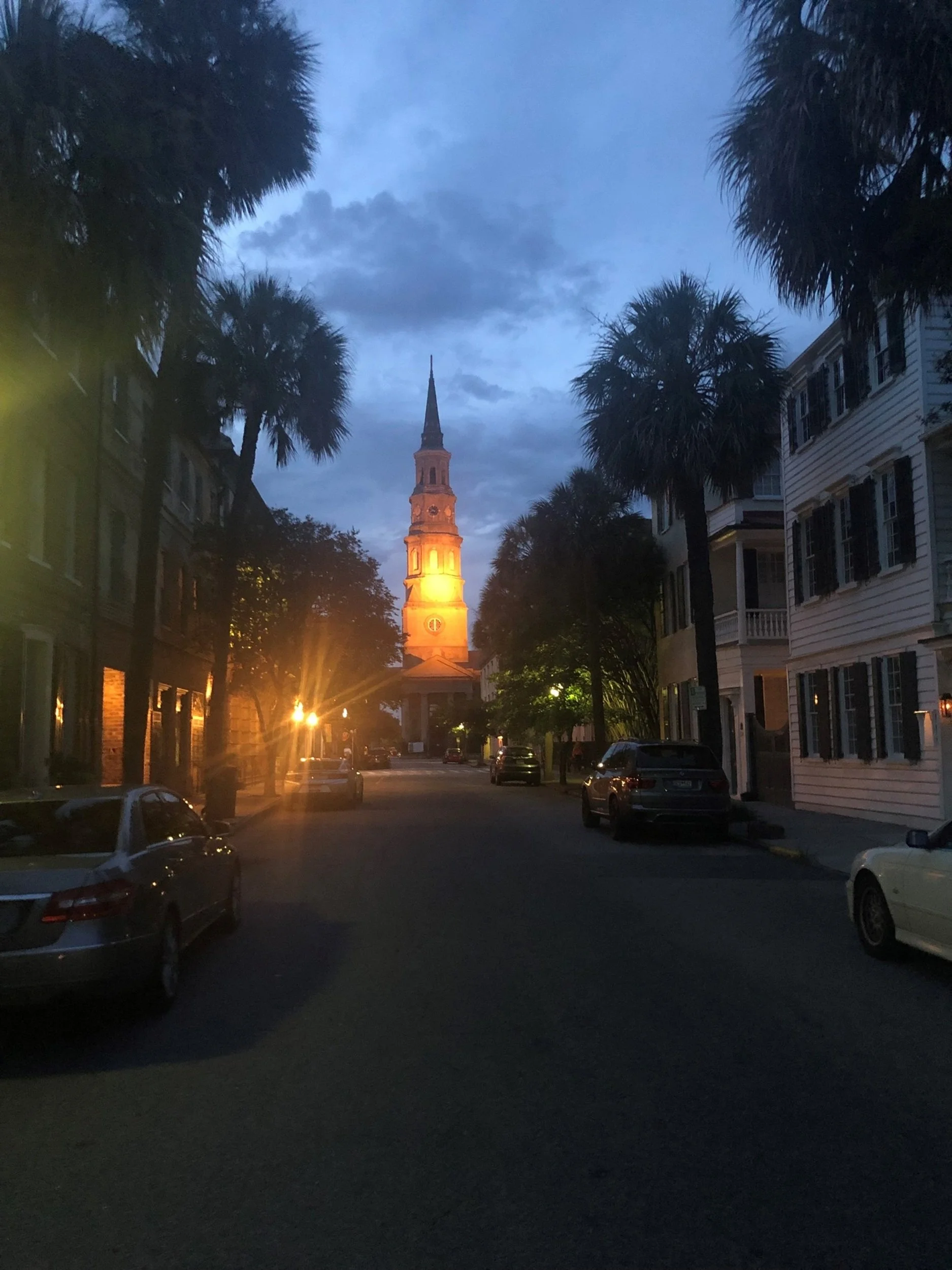 Sunset over a street with palm trees, historic church with illuminated steeple, and parked cars along the road.