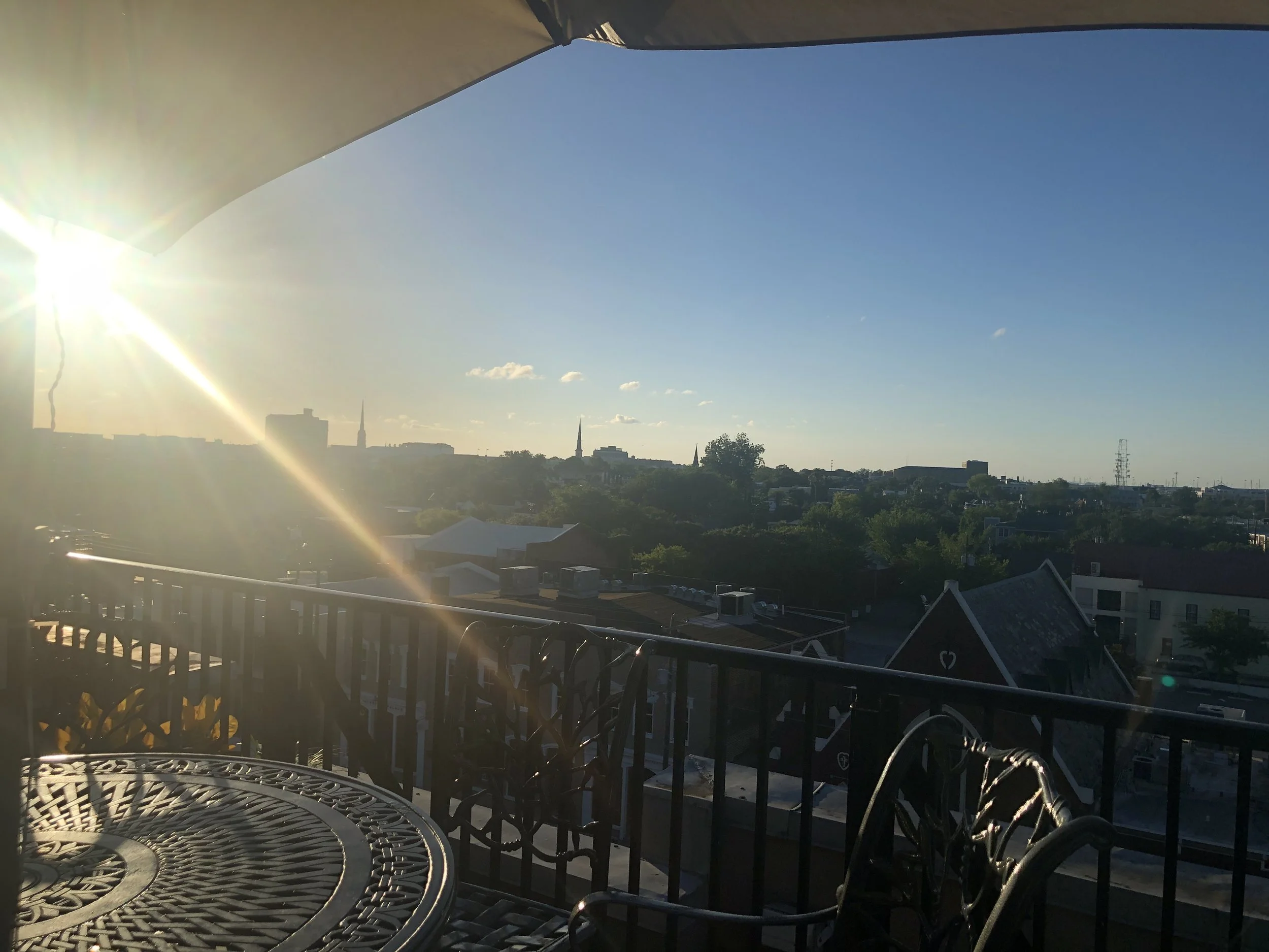 Charleston Evening (Rooftop View): Golden afternoon light stretches across Charleston's historic skyline from a rooftop terrace, where timeless architecture and open skies create a quiet sense of anticipation. The elevated perspective captures both t