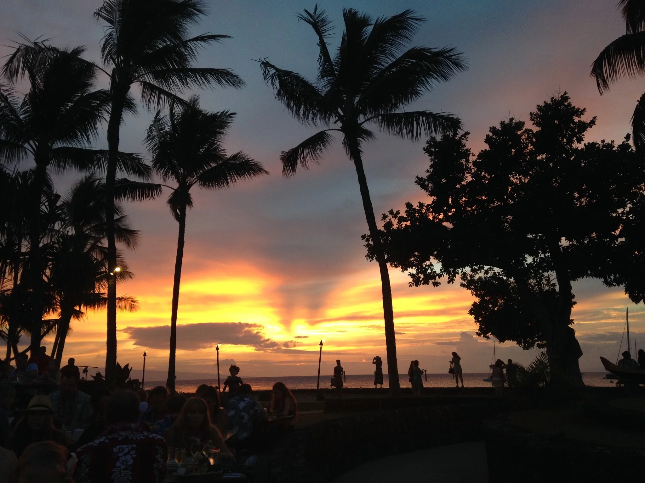 Aloha Evening: As the Hawaiian sun slips below the Pacific, silhouettes of swaying palms, evening diners, and strolling guests create a scene that captures the warmth and rhythm of island hospitality. Rich golden light and dramatic tropical skies tra
